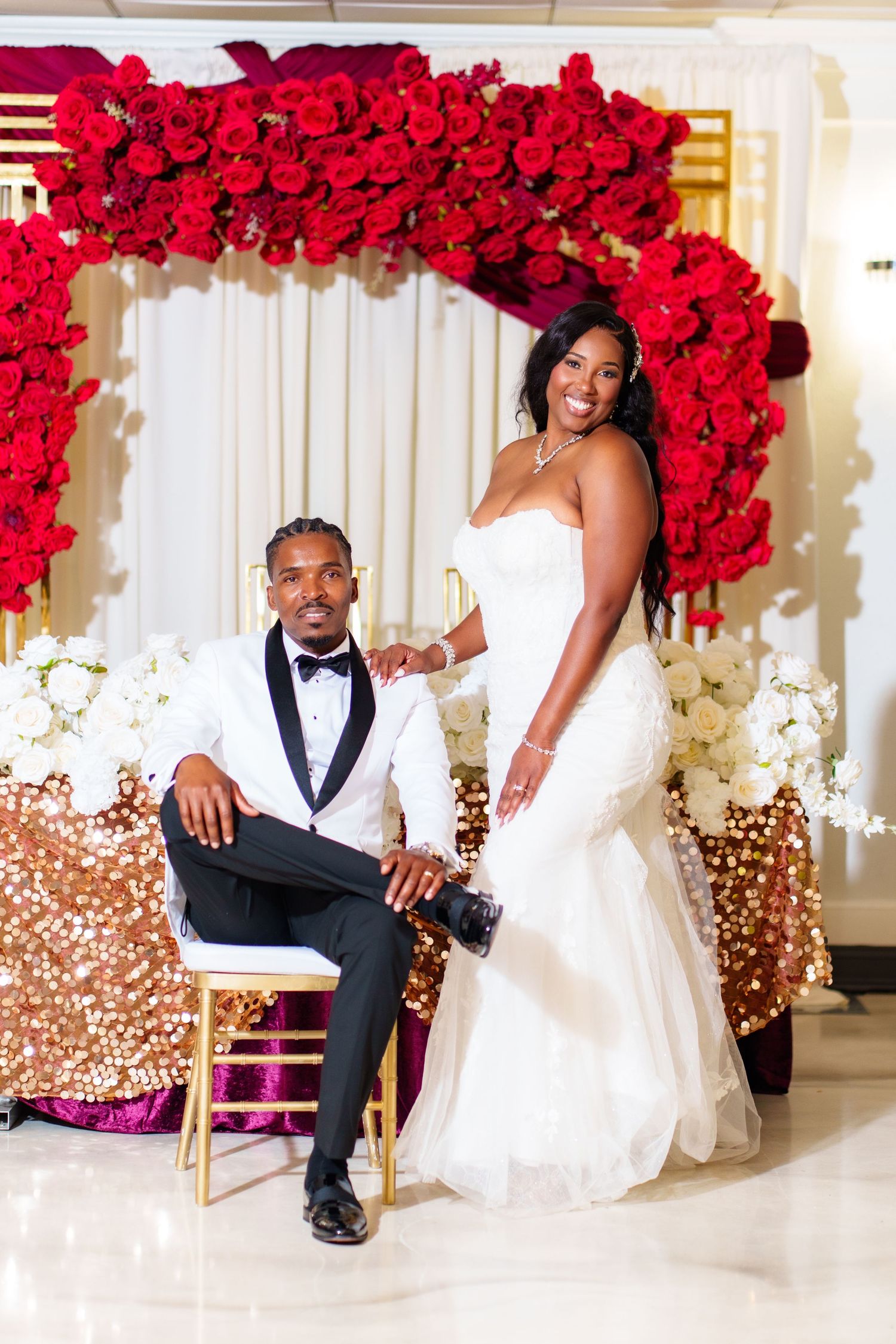 Bride in white strapless gown and groom in tuxedo pose before red rose arch with gold sequin tablecloth at wedding reception.