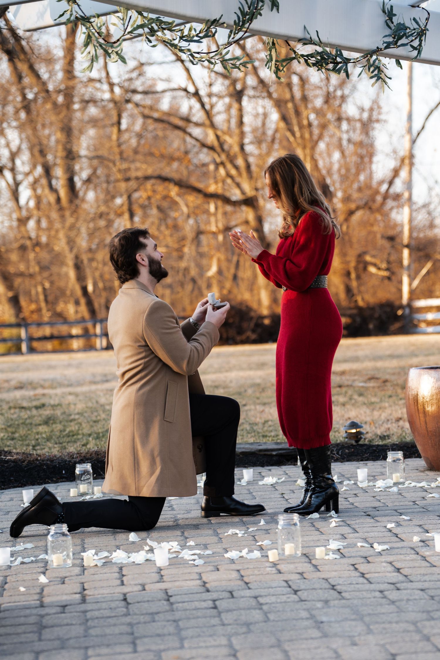 Man on one knee proposing to woman in red dress outdoors surrounded by candles and rose petals.