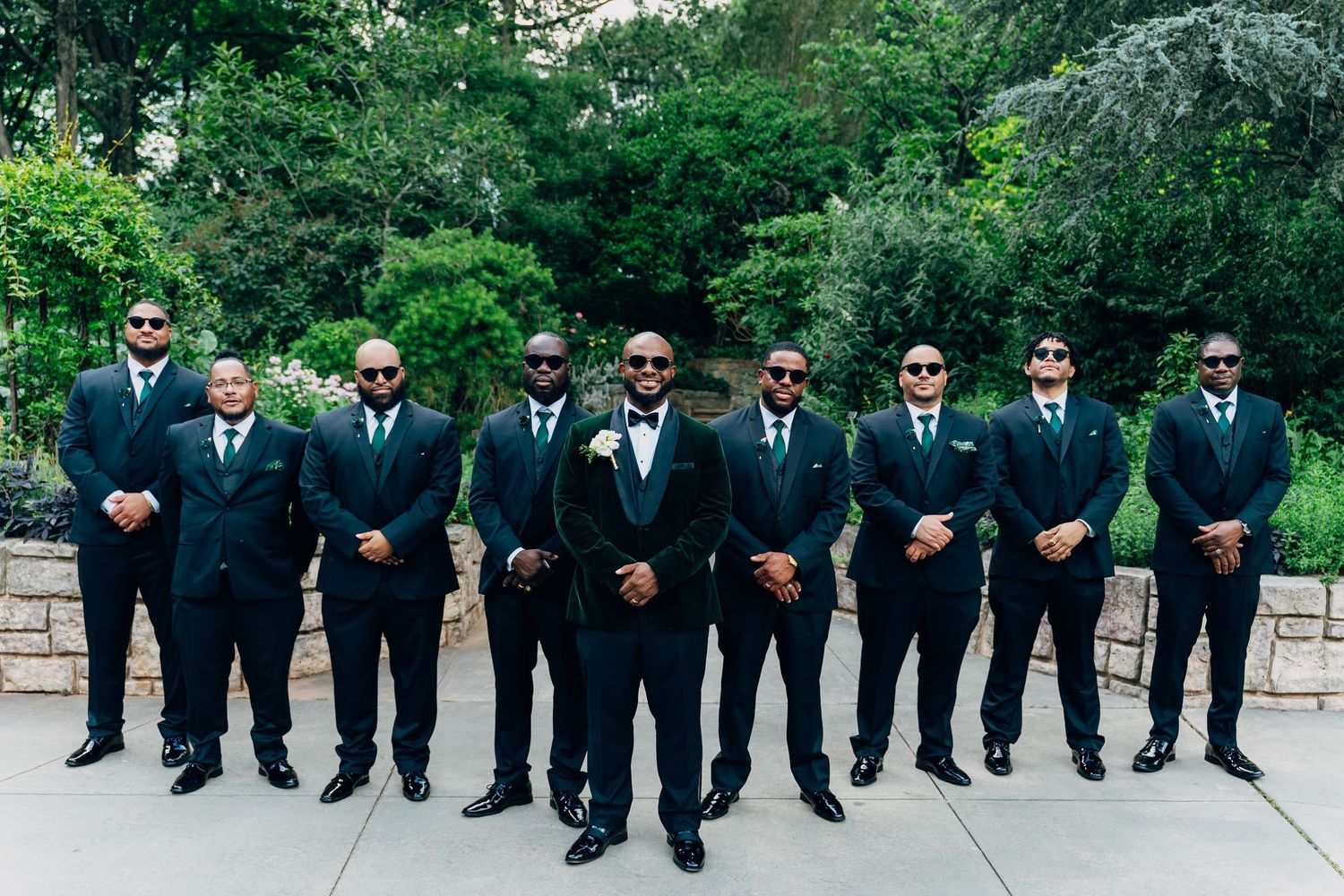 Groom and groomsmen in matching black suits and sunglasses posing outdoors at a wedding.