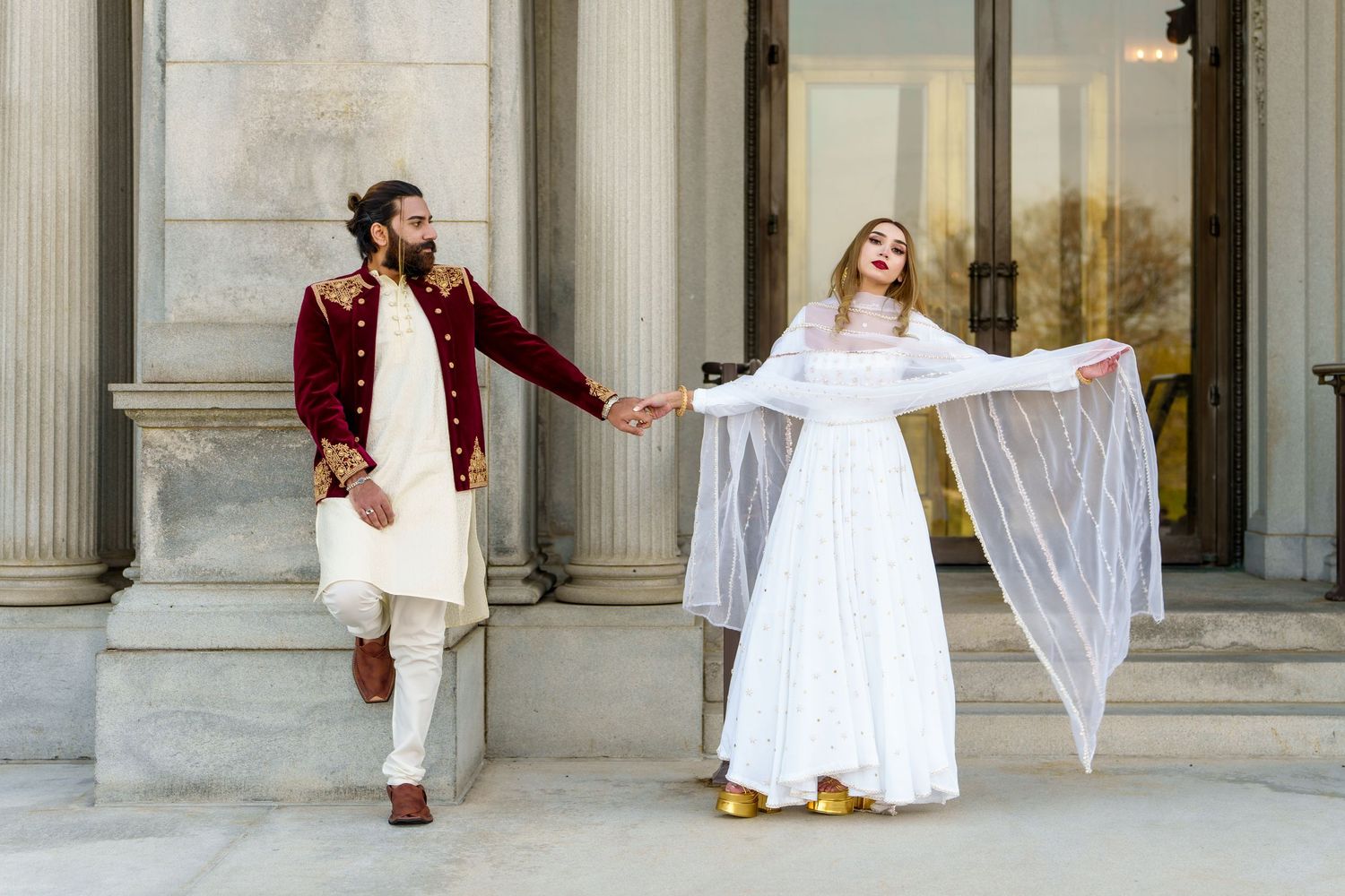 Couple in traditional South Asian wedding outfits posing outside classical stone columns.