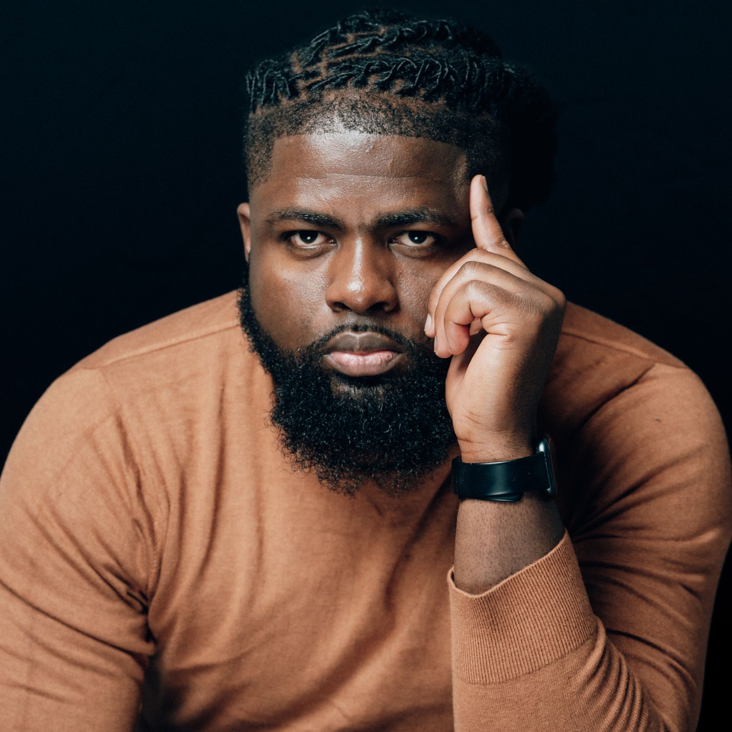 Confident bearded Black man in tan sweater poses with finger to temple against dark studio background.