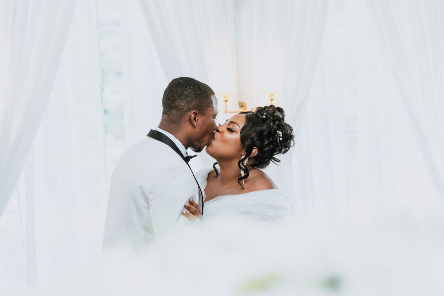 Romantic Black couple sharing a tender kiss framed by sheer white curtains at wedding.