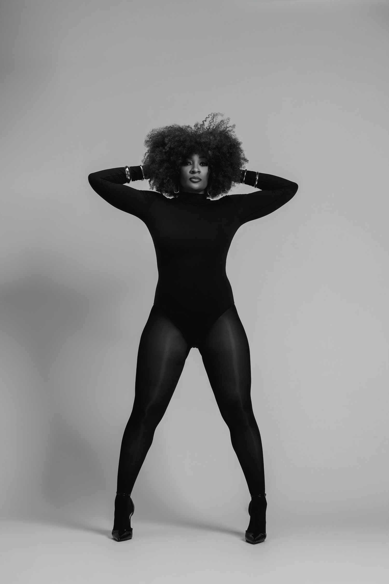 Black and white photo of a woman in a black bodysuit and tights, posing confidently with hands behind her afro hair.