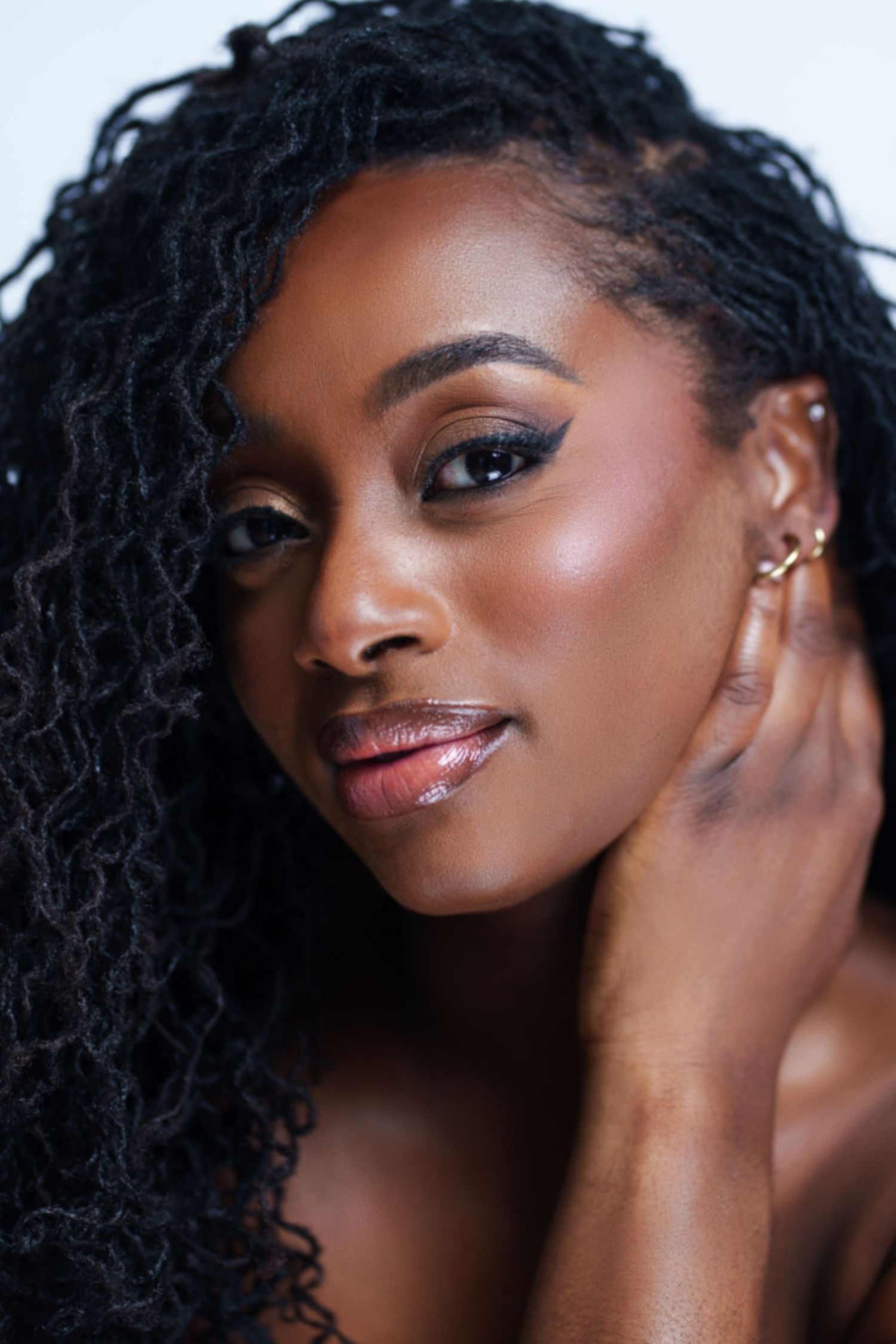 A young Black woman with long locs poses gracefully, wearing gold ear cuffs and subtle glam makeup.