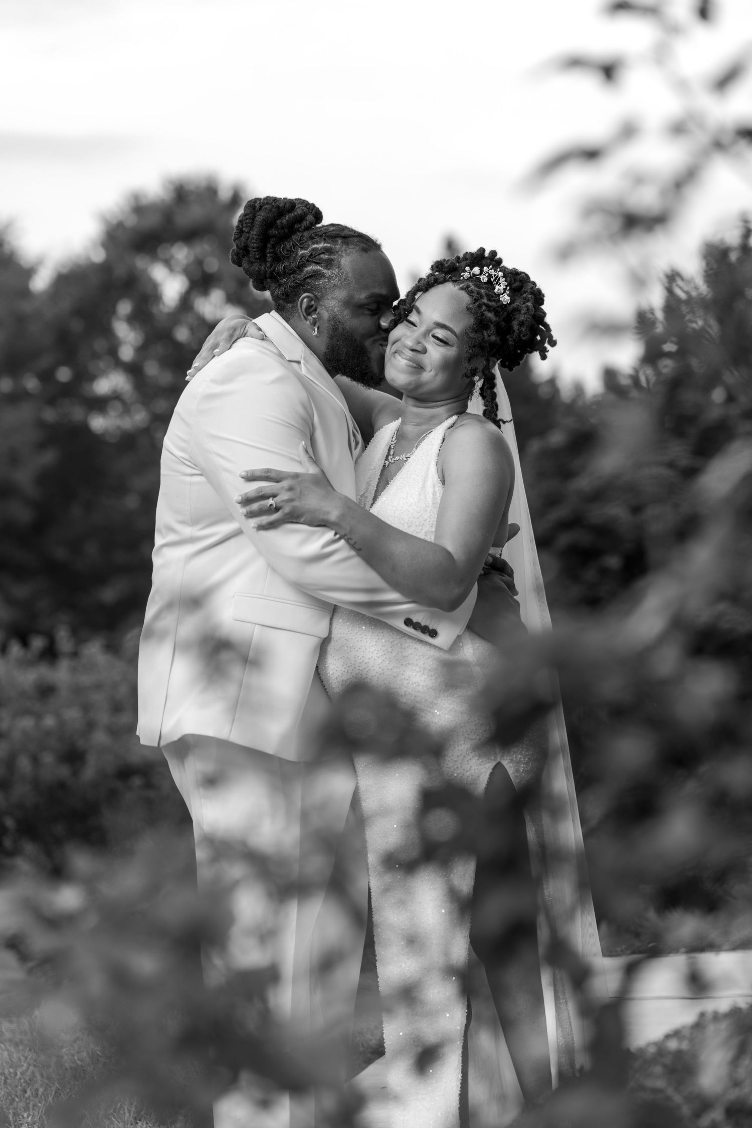 Black and white photo of two brides embracing and smiling on their wedding day outdoors among lush greenery.