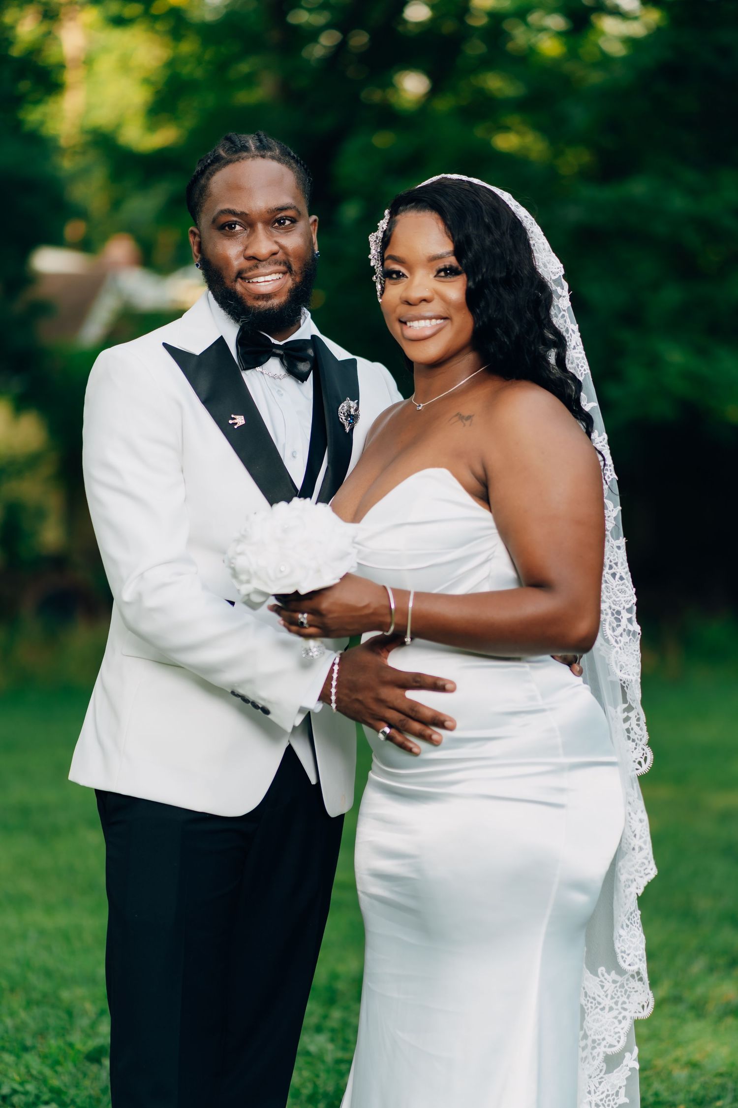 A smiling bride and groom pose outdoors, she in a white gown with veil, he in a white tuxedo jacket.
