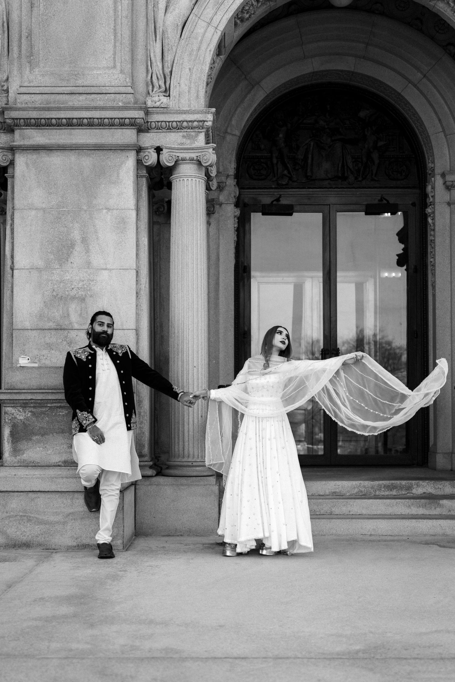 Black and white photo of a South Asian bride and groom holding hands before a grand archway.