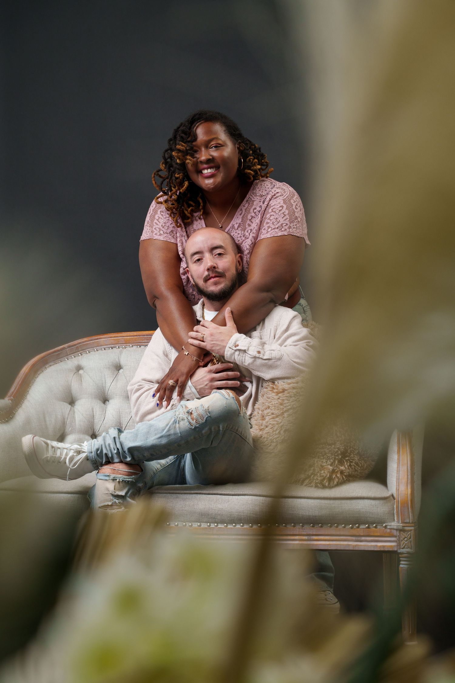 A couple shares a tender embrace on a tufted white sofa, the woman hugging the man from behind, both smiling warmly.