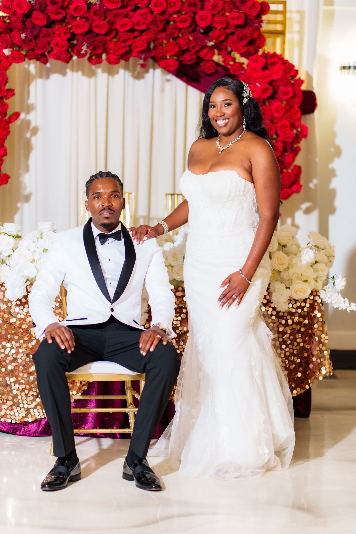 Bride in white strapless gown stands beside groom in white tuxedo jacket before red rose floral arch.