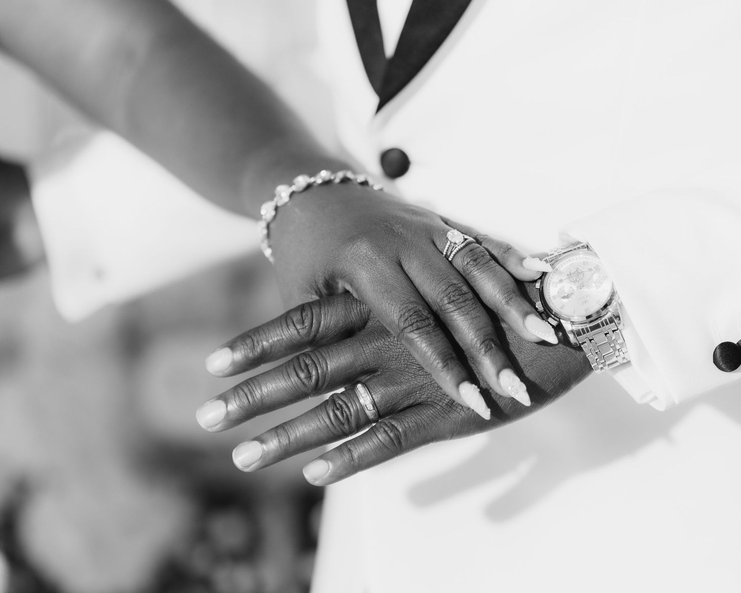 Black and white photo of a couple holding hands, showing wedding rings, a bracelet, and a luxury watch.