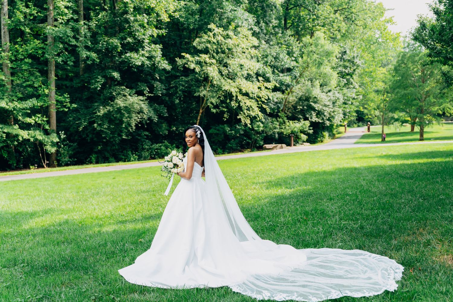 Bride in elegant white ball gown with long veil holding bouquet on lush green lawn surrounded by trees.
