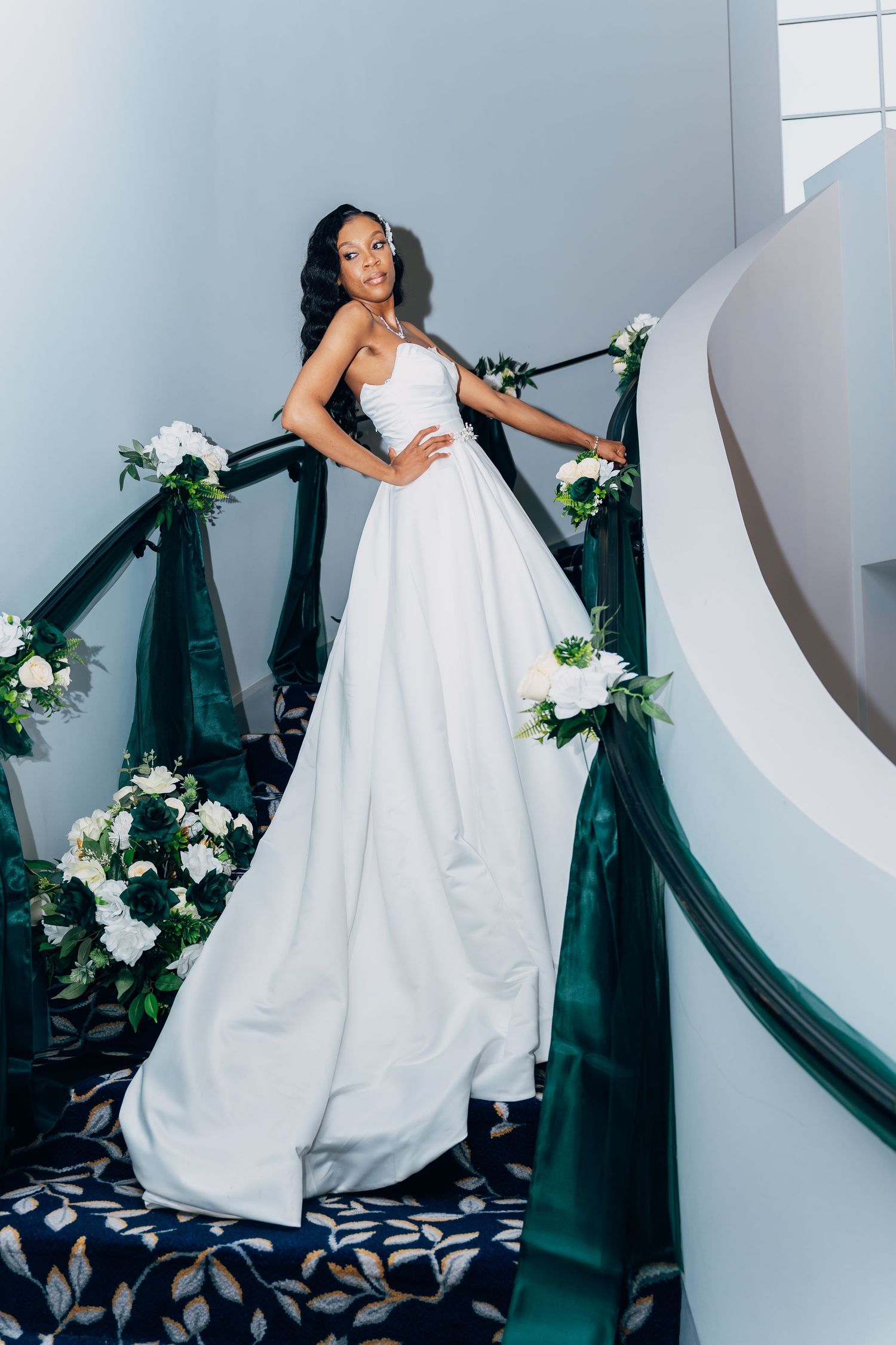 Elegant bride in white gown poses on spiral staircase adorned with green ribbon and white floral bouquets.