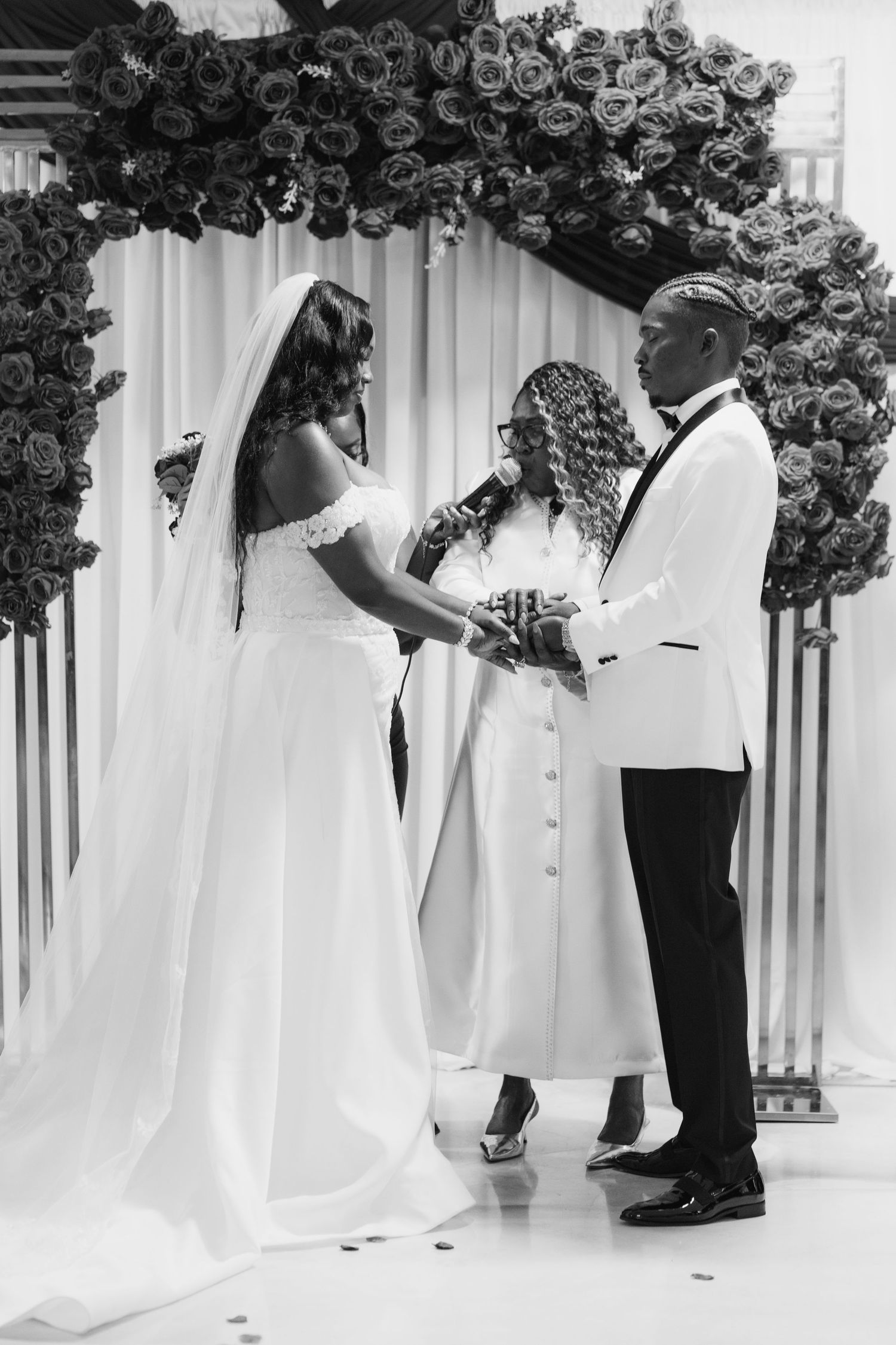 A bride and groom exchange vows before an officiant under a floral arch in black and white.