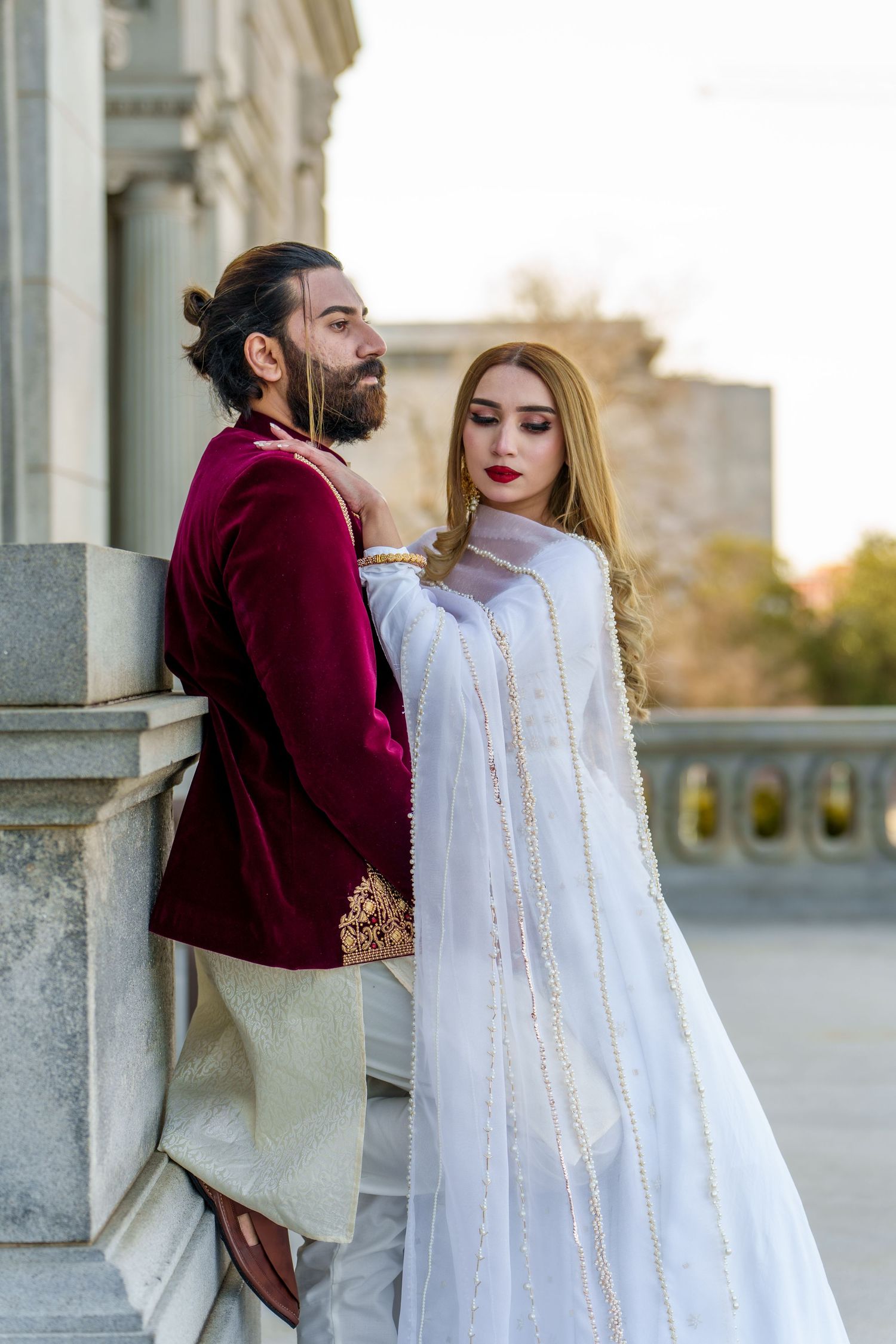 Couple in elegant traditional South Asian wedding attire posing by stone pillar.