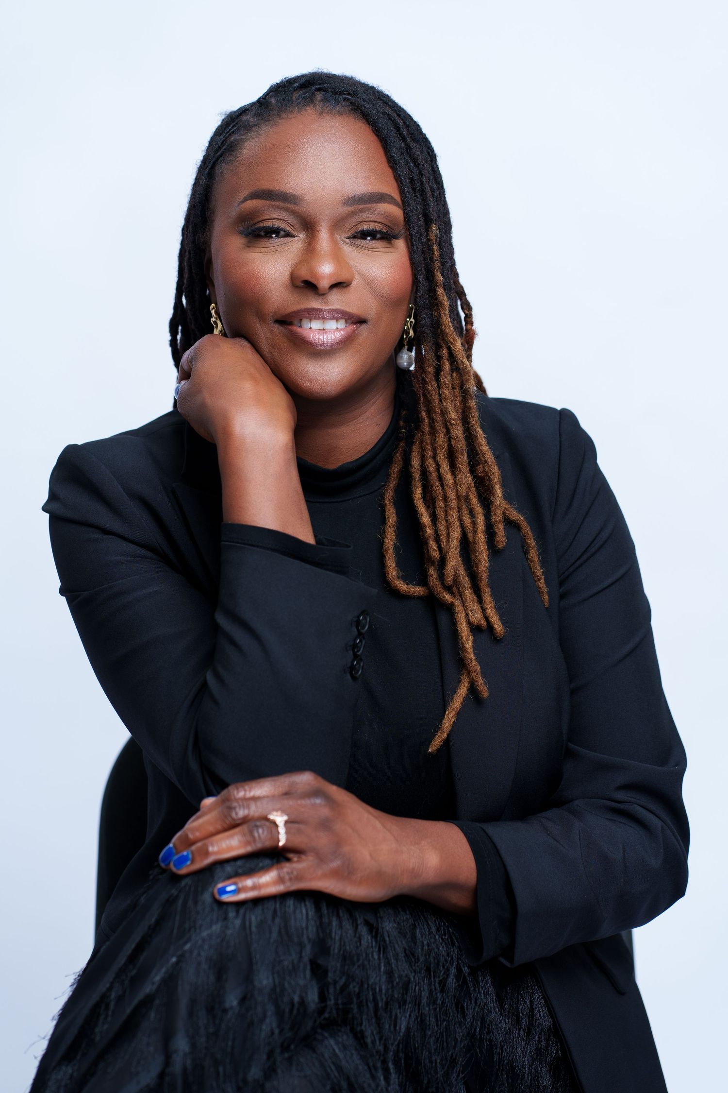 Professional portrait of a smiling woman with locs wearing a black blazer, posing confidently against a white background.