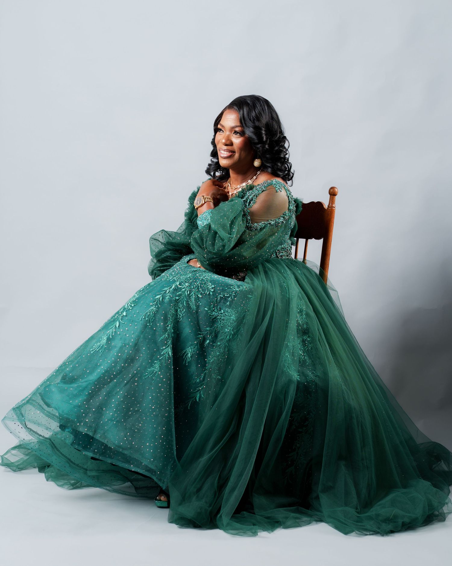 Woman in elegant emerald green ball gown with sequins and tulle, seated gracefully, smiling, wearing pearl earrings.
