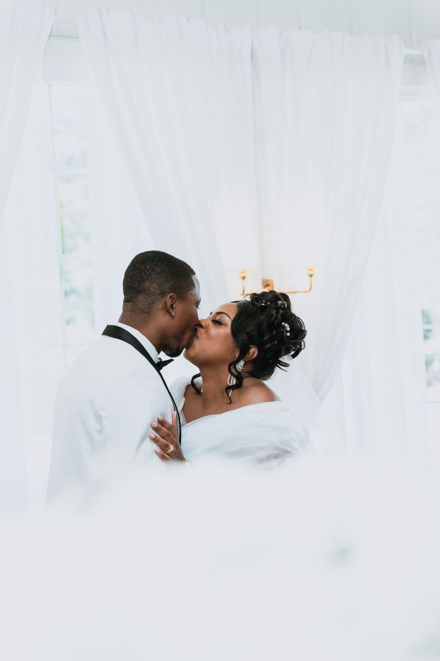 Bride and groom sharing a romantic kiss framed by white sheer curtains at their elegant wedding venue.