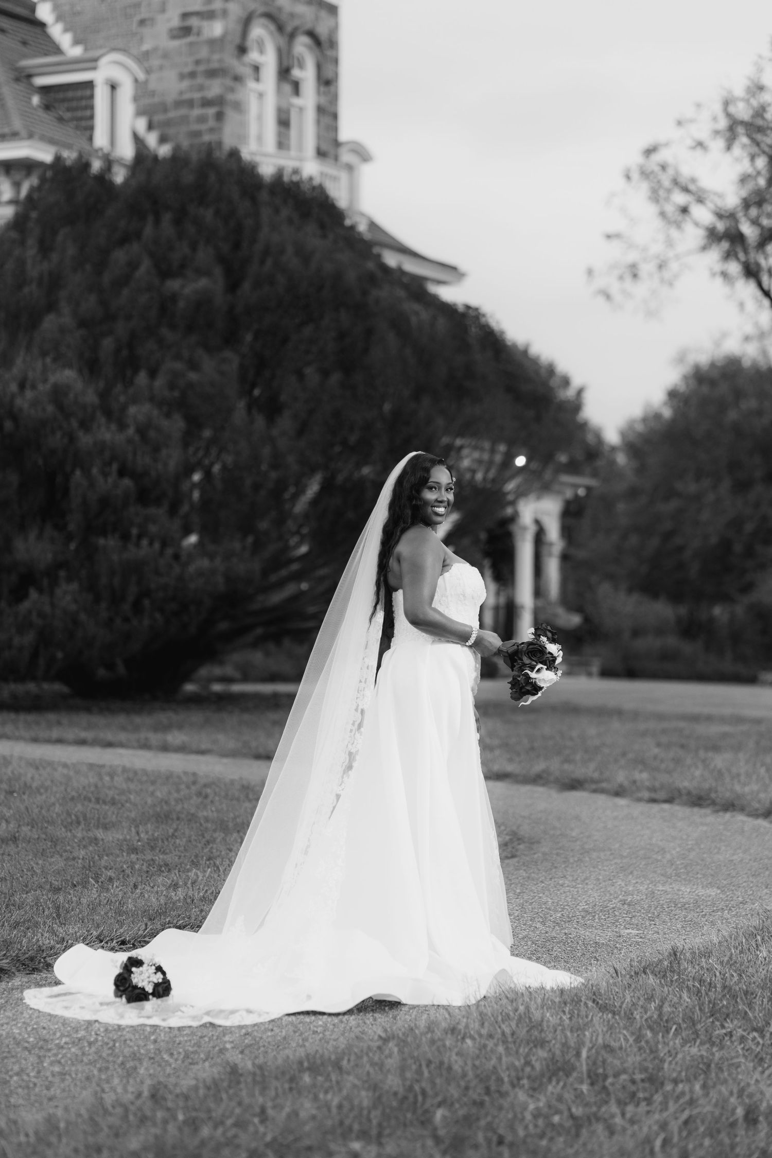 Black and white bridal portrait of a smiling bride in a flowing gown and long veil holding a bouquet outdoors.