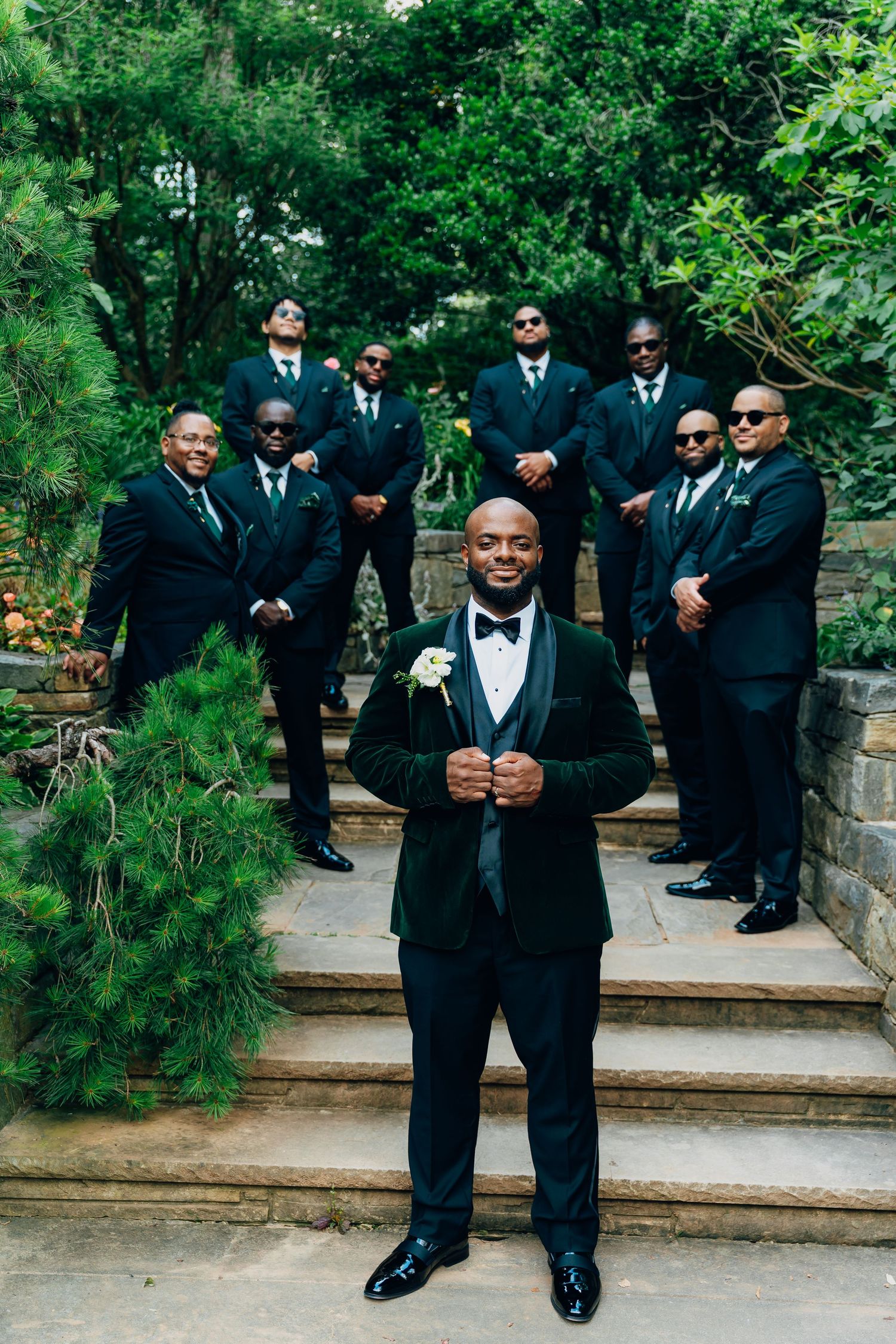 Groom in green velvet tuxedo poses with groomsmen in black suits on stone steps surrounded by lush greenery.