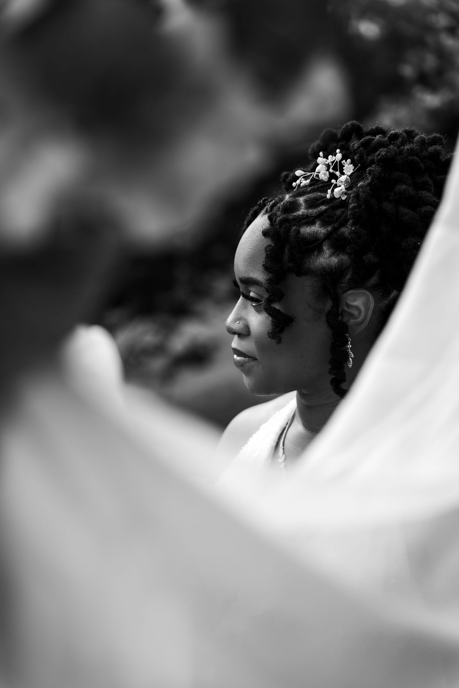 Black and white portrait of a bride with floral hair accessories and locs, gazing softly through a flowing veil.