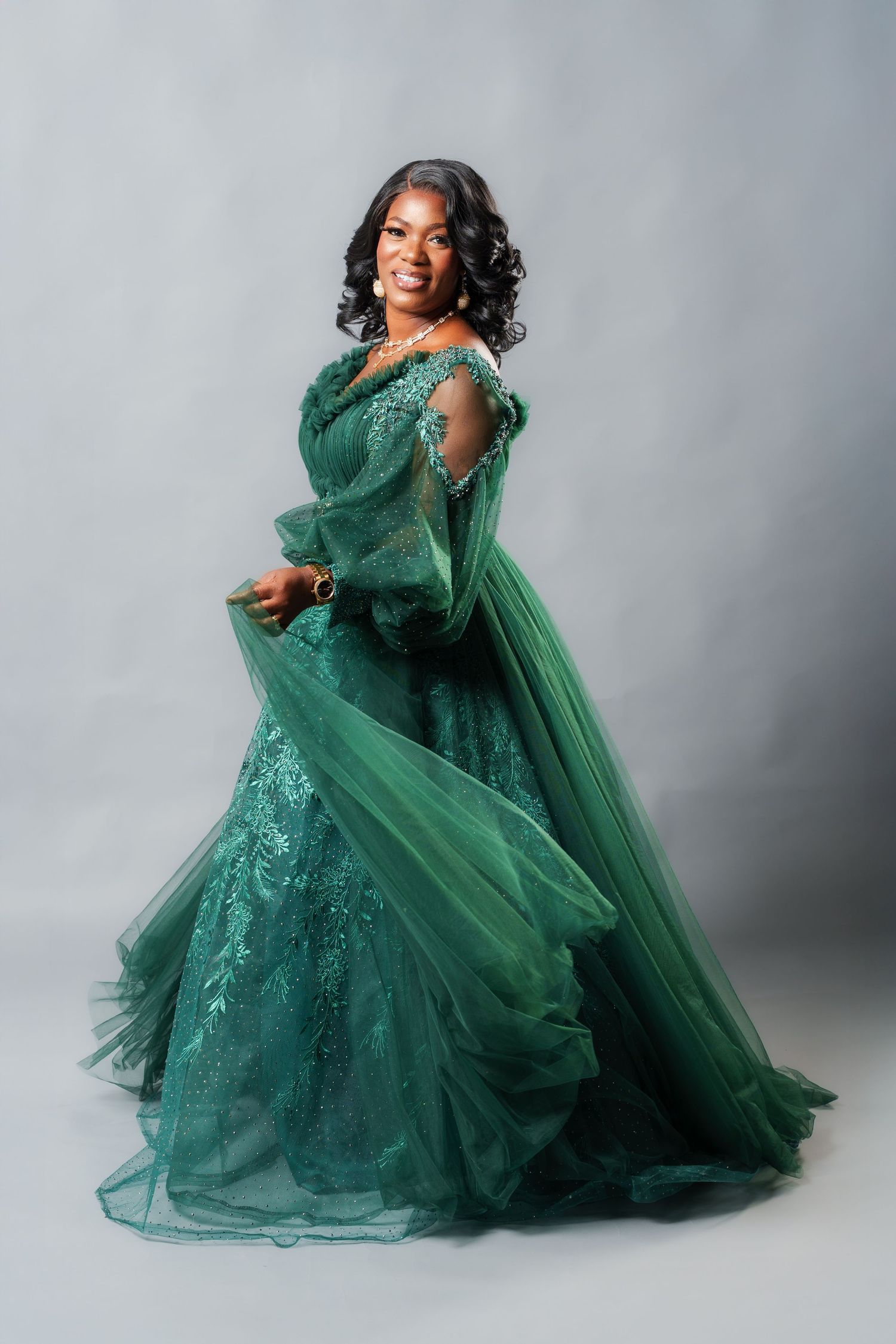 Woman in elegant emerald green tulle ball gown smiling, posing gracefully against gray studio background.