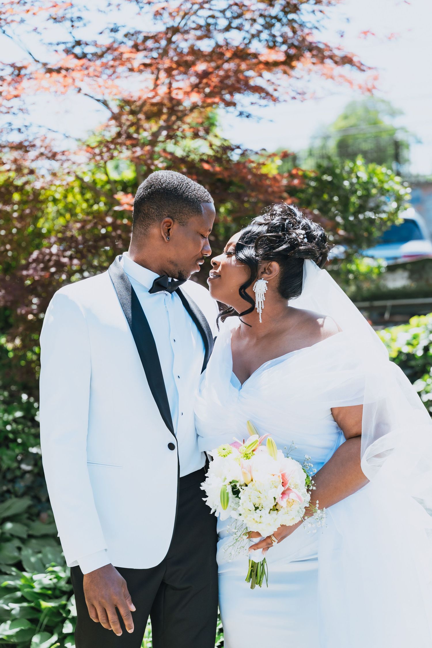 A bride and groom share a romantic moment outdoors, with the bride holding a white bouquet and the groom in a white tuxedo.