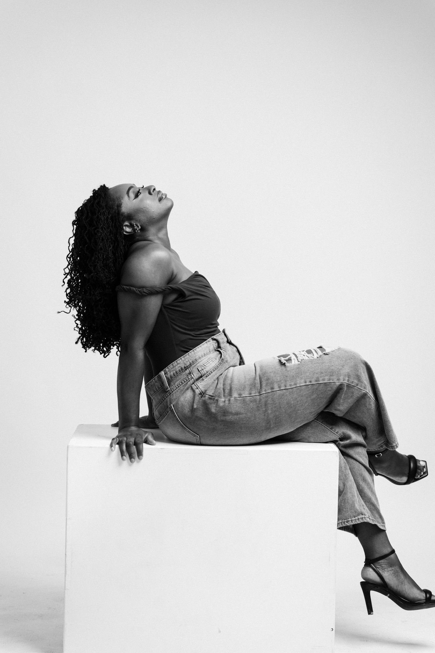 Black and white photo of a woman in jeans and heels posing on a white cube with head tilted back.