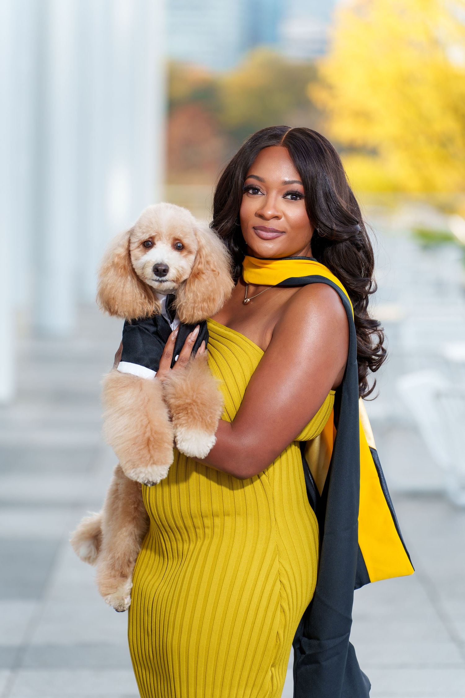 A woman in a yellow dress holds an apricot poodle wearing a black outfit while posing outdoors in autumn.