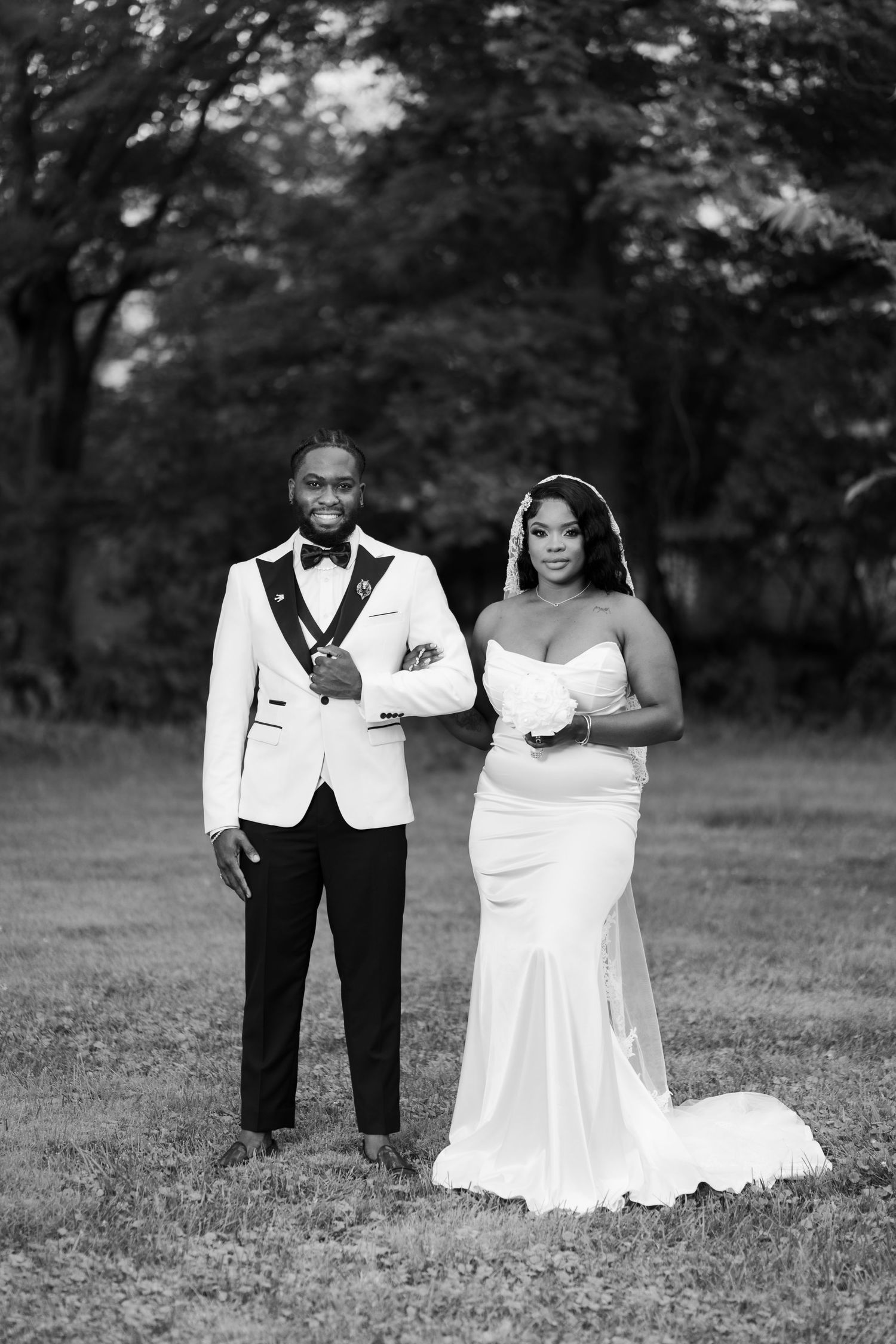 Black and white wedding photo of a couple in formal attire, groom in white tuxedo and bride in fitted gown, outdoors.