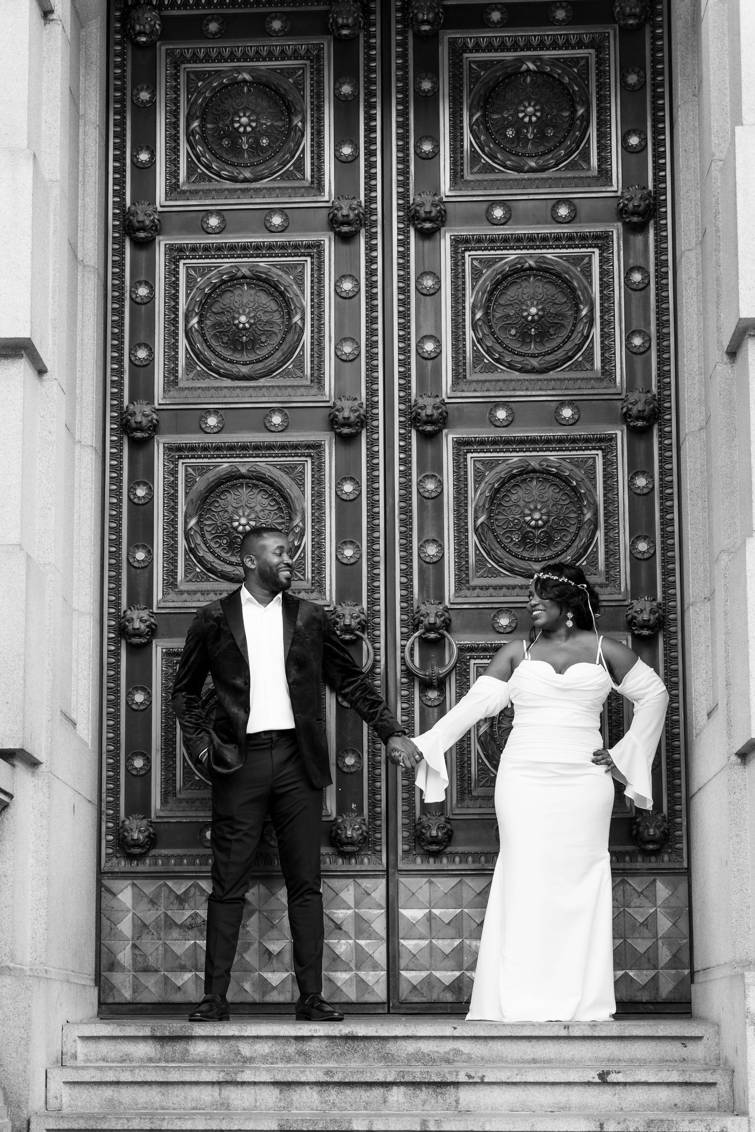 Black and white photo of a bride and groom holding hands before ornate cathedral doors on stone steps.