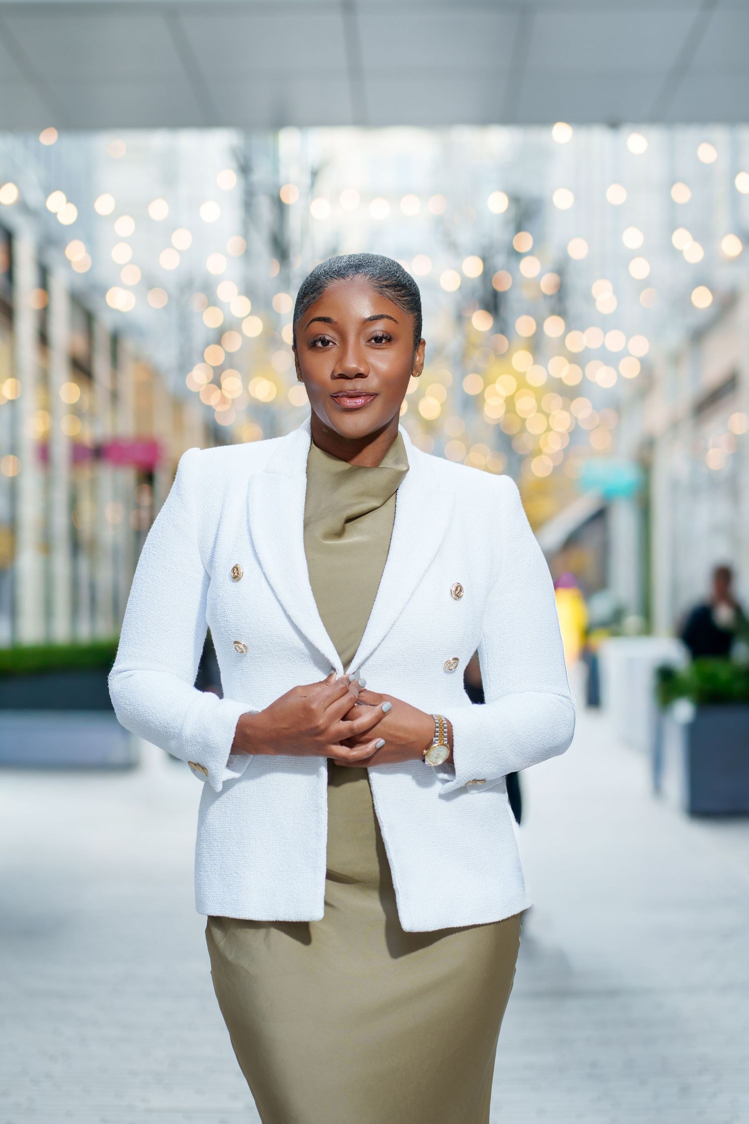 A professional woman in a white blazer and olive dress stands in a corridor with bokeh lights behind her.