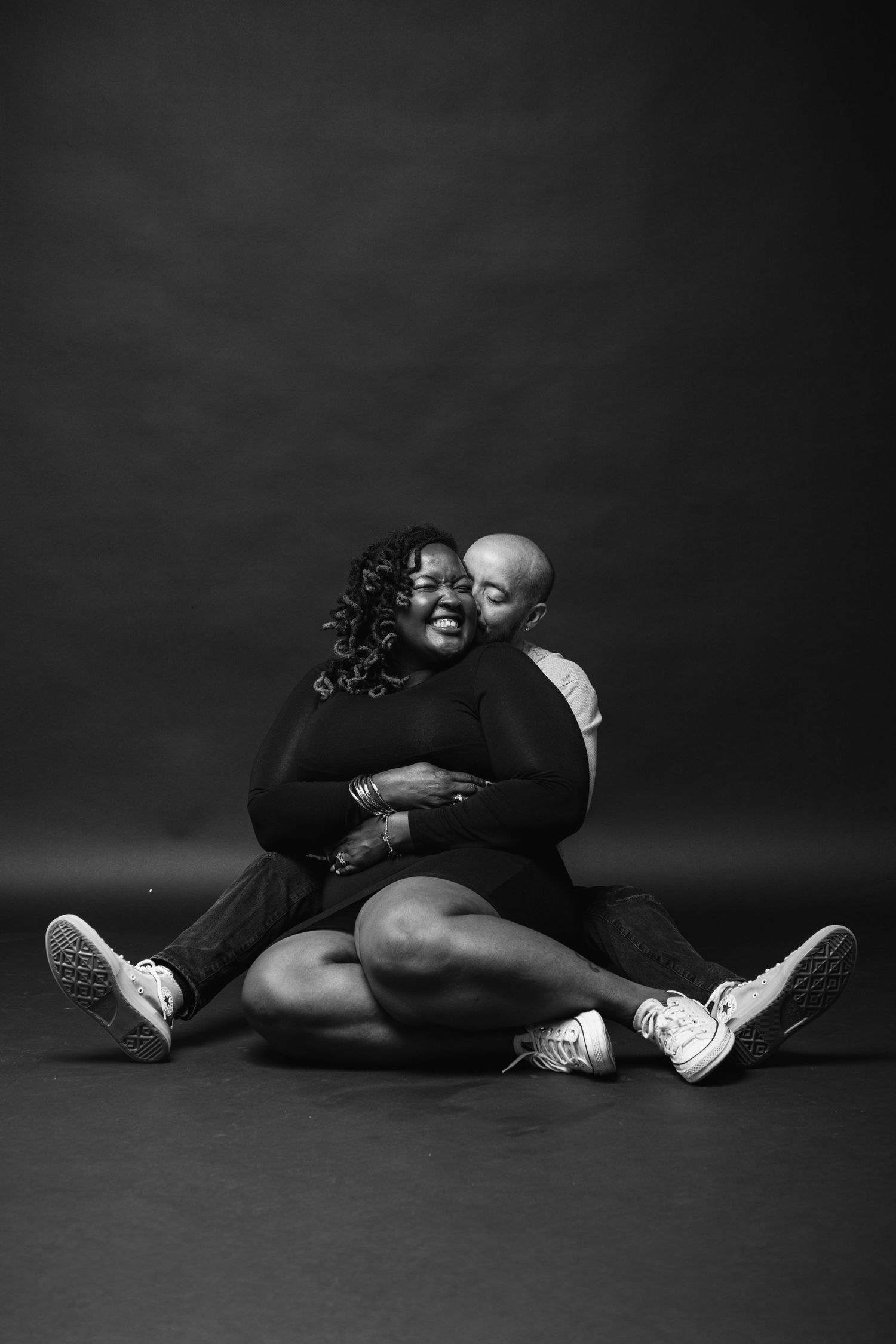 Black and white photo of a couple sitting on floor, man embracing woman from behind, both wearing sneakers.