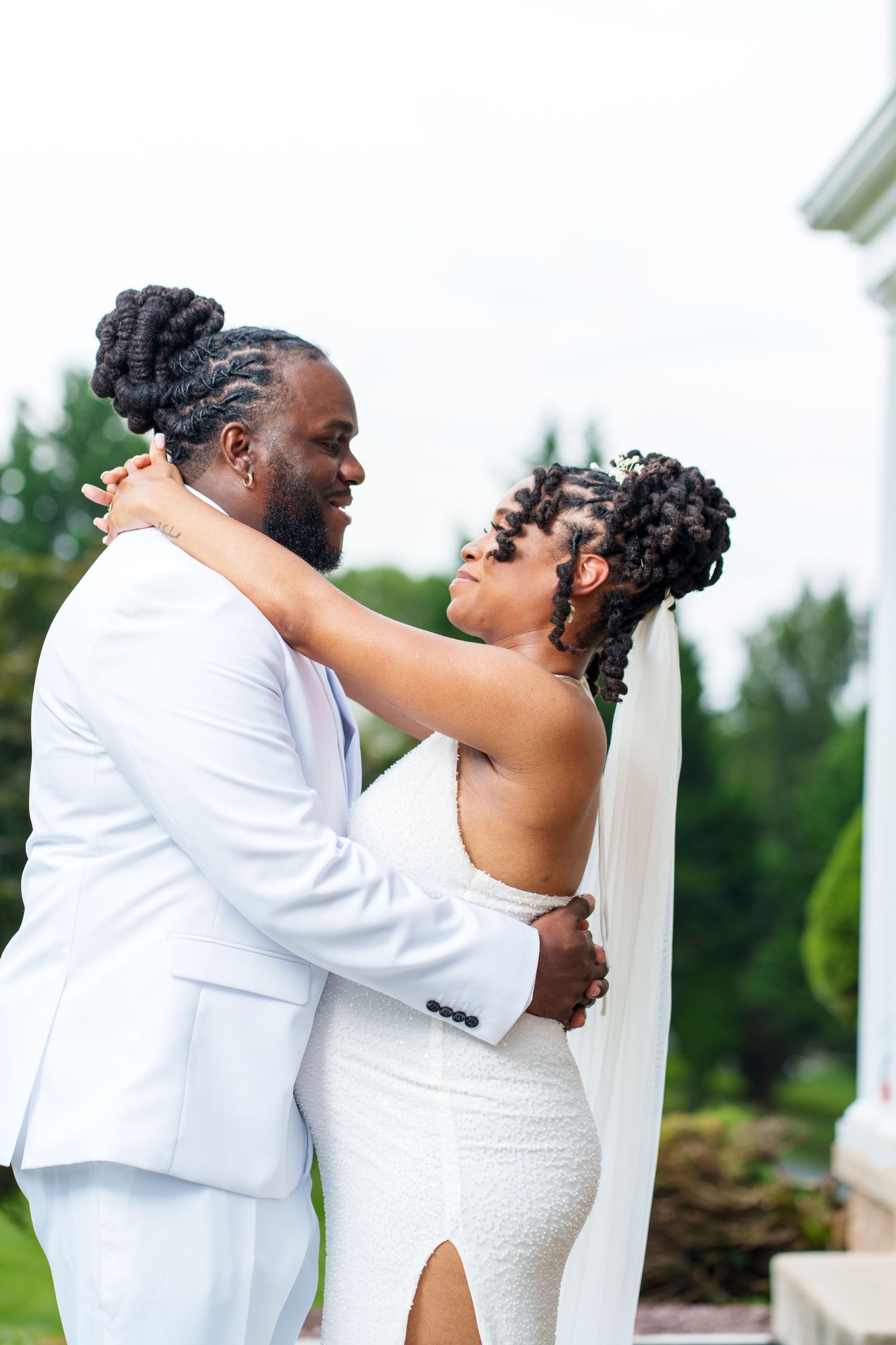 A Black bride and groom in white wedding attire embrace outdoors, sharing a romantic moment surrounded by lush greenery.