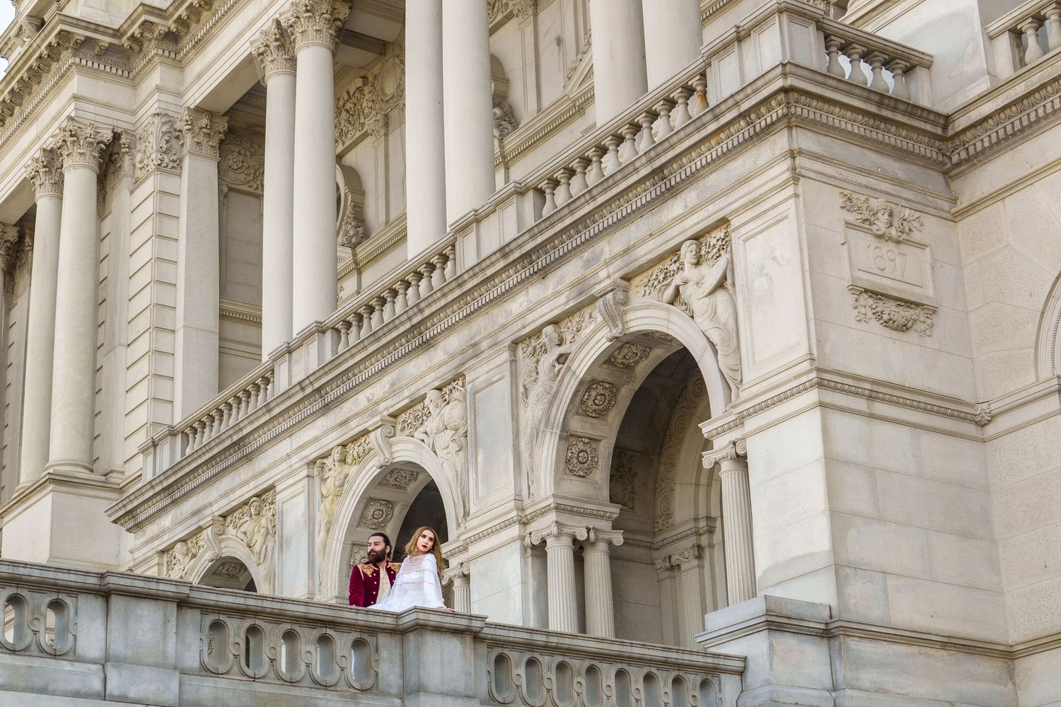 Couple in traditional wedding attire standing on balcony of grand classical building.