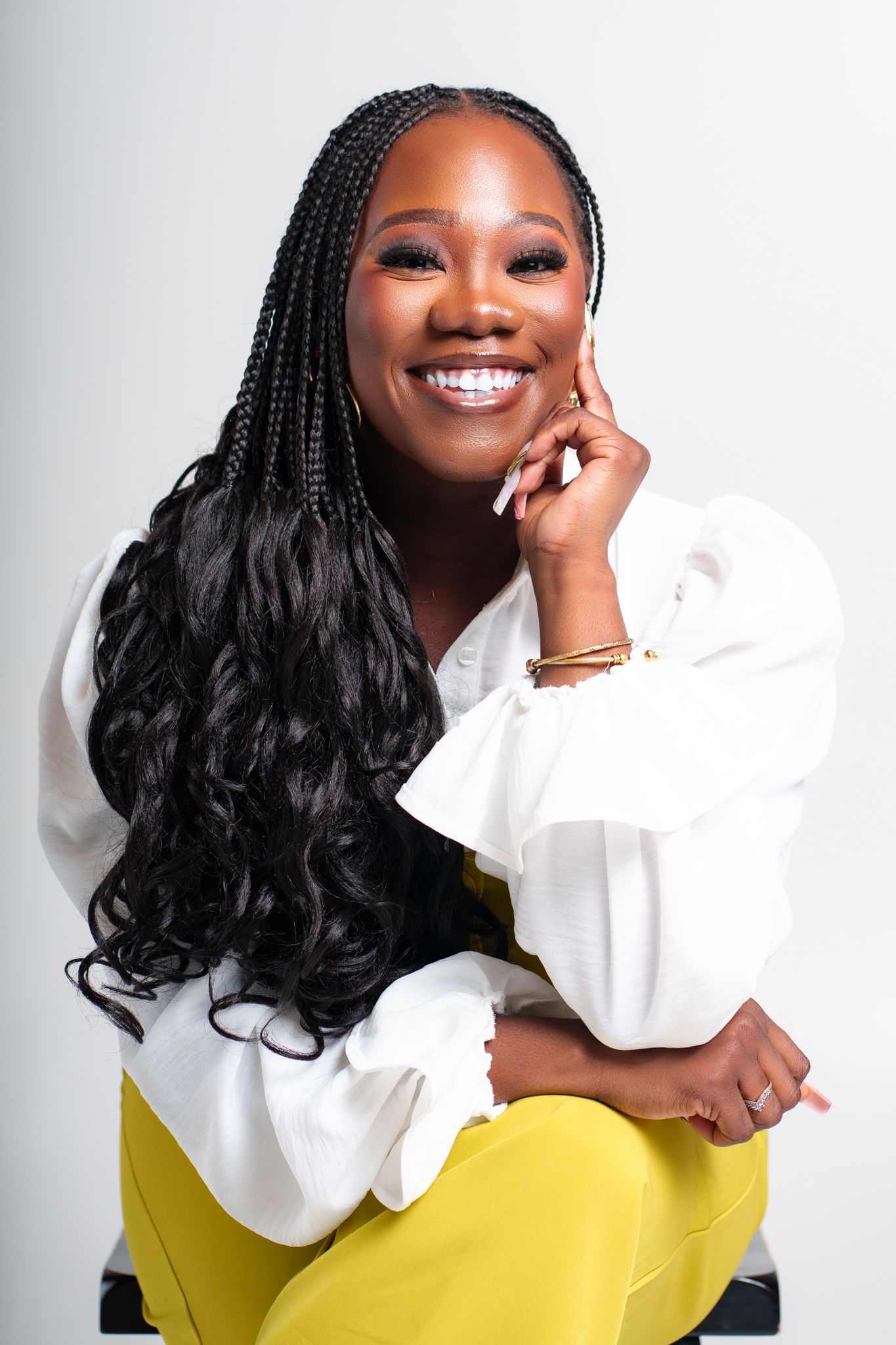 A smiling woman with long box braids sits wearing a white ruffled blouse and yellow pants on a white background.