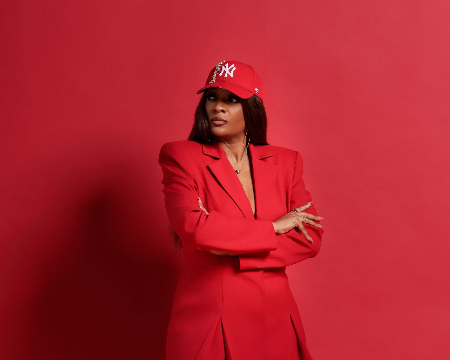 A confident woman in an all-red suit and NY Yankees cap poses with arms crossed against a red background.