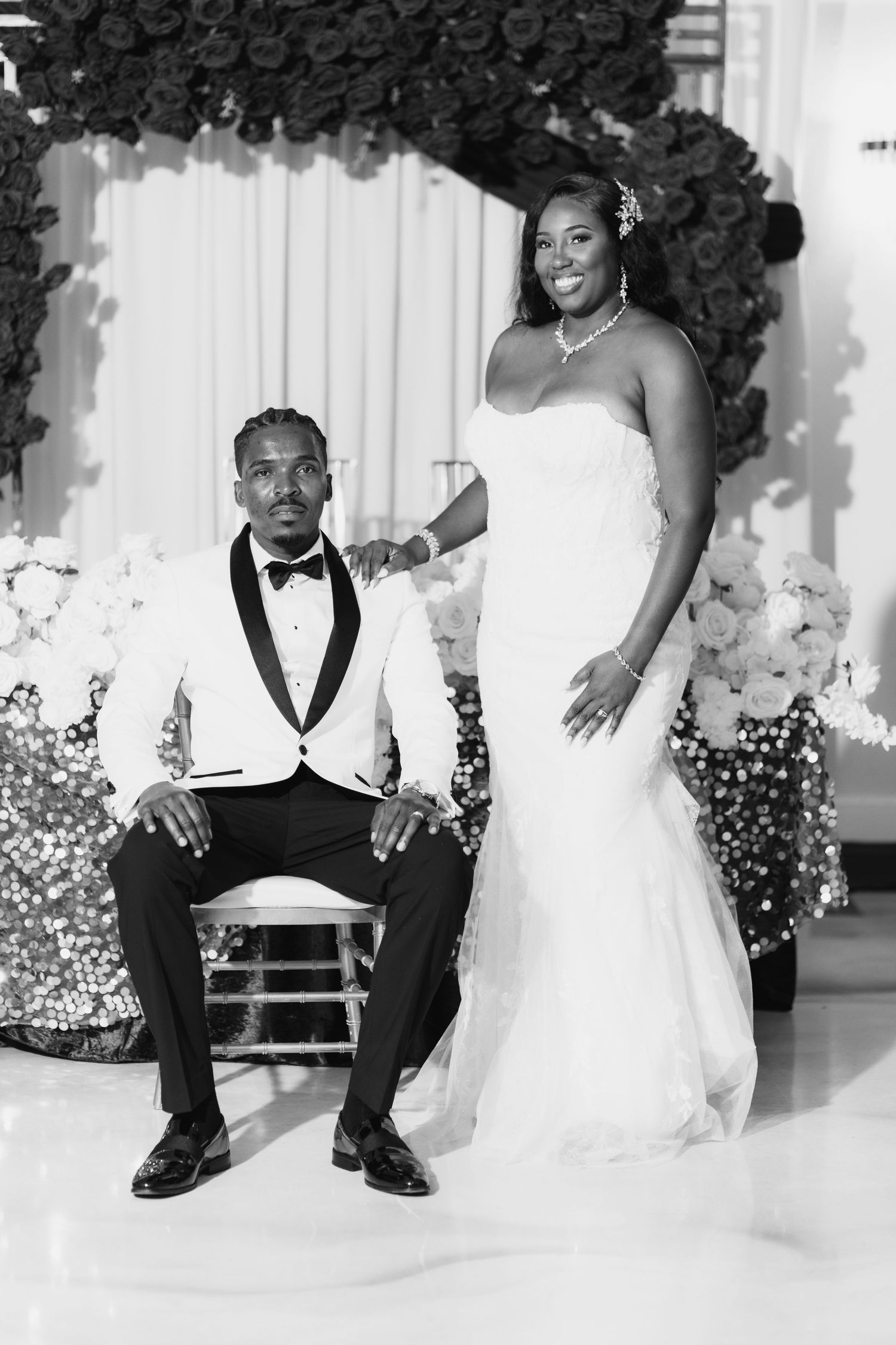 Black and white wedding photo of bride in mermaid gown and groom in tuxedo posing elegantly before floral backdrop.
