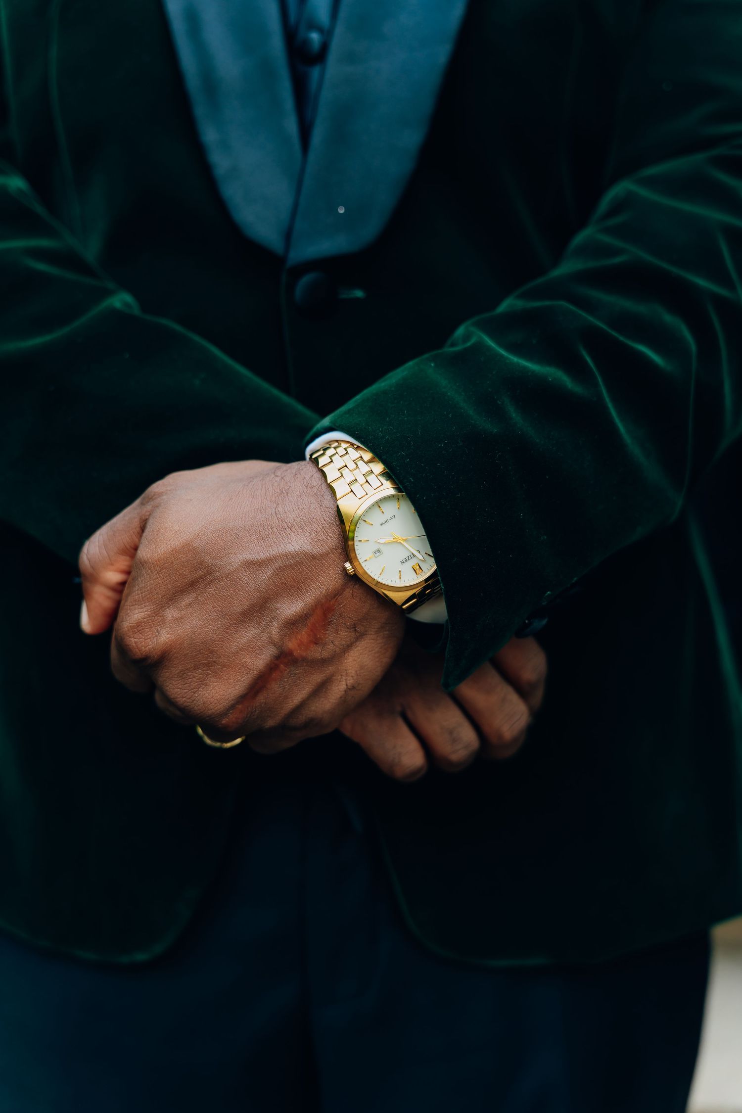 Man in green velvet blazer clasping hands, wearing a gold wristwatch and ring, close-up detail shot.