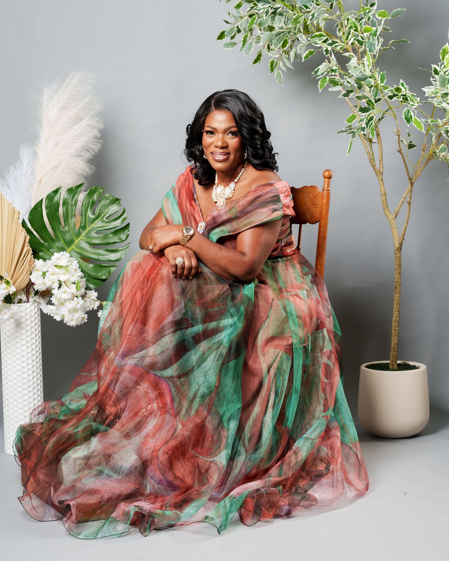 Woman in colorful pink and green floral gown seated in chair with tropical plants in studio setting.