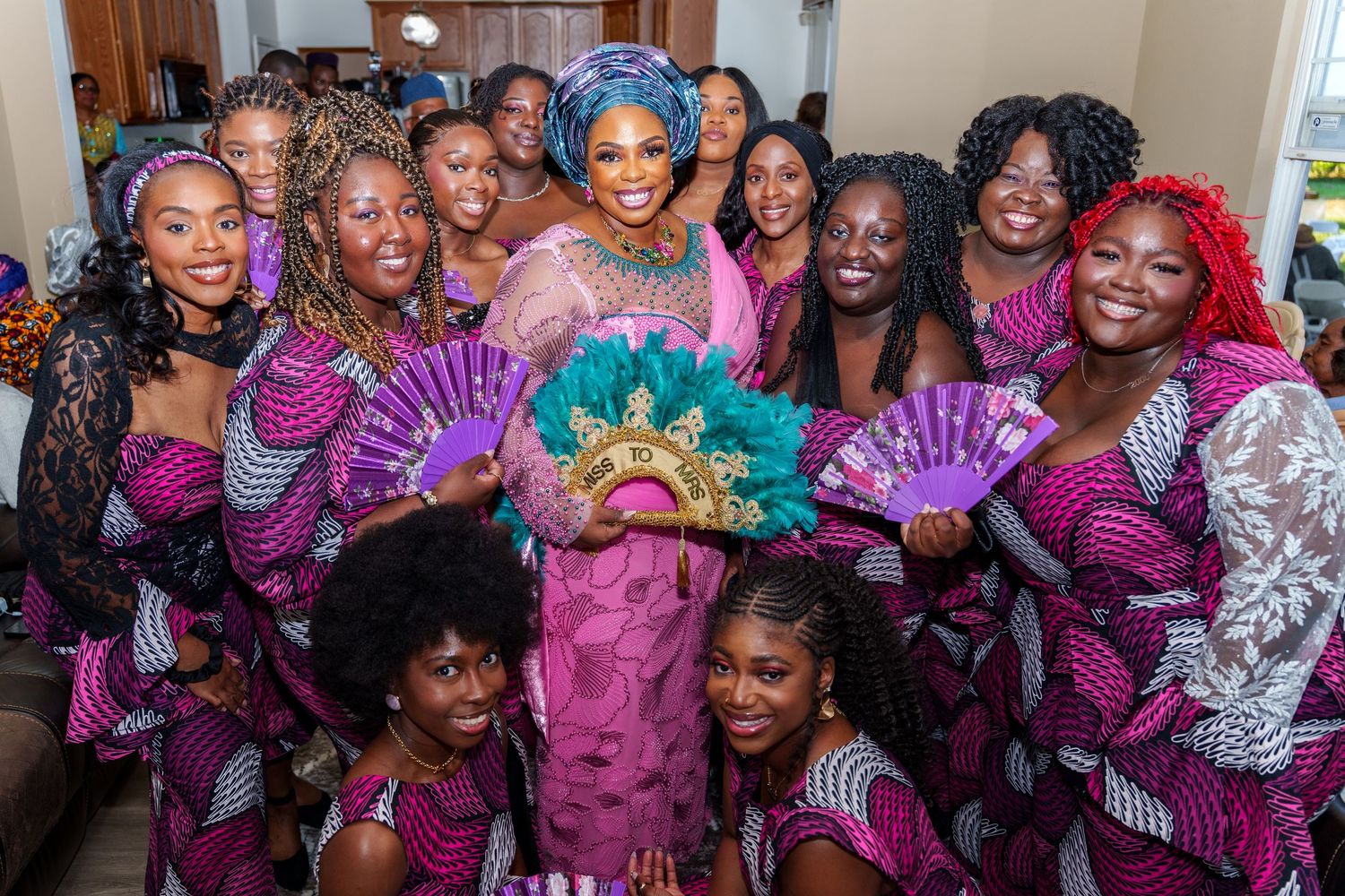 A bride in pink lace and teal headpiece poses with bridesmaids in purple Ankara print dresses holding teal fans.