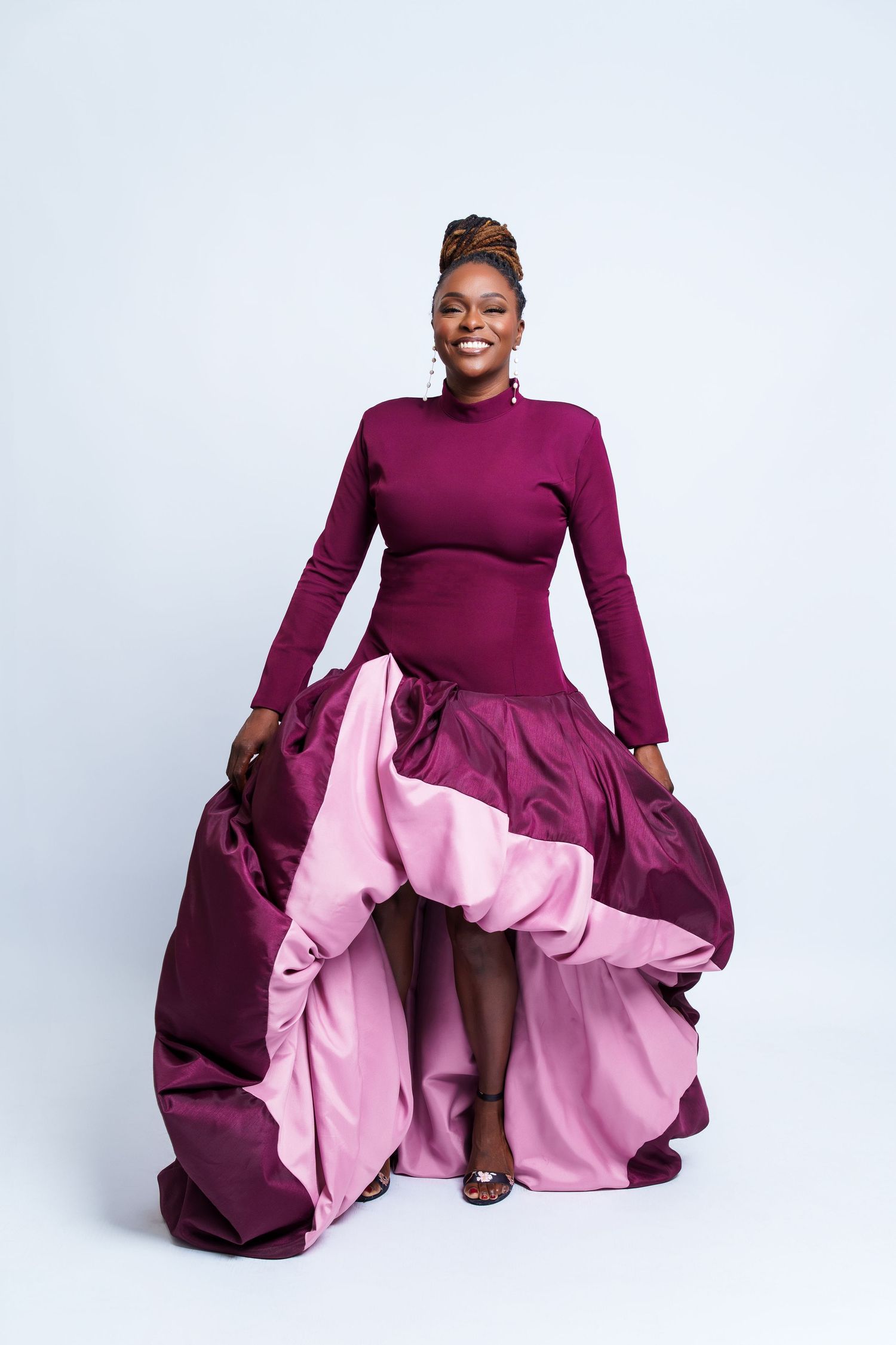 Woman in elegant burgundy and pink ballgown smiling confidently against light blue background.