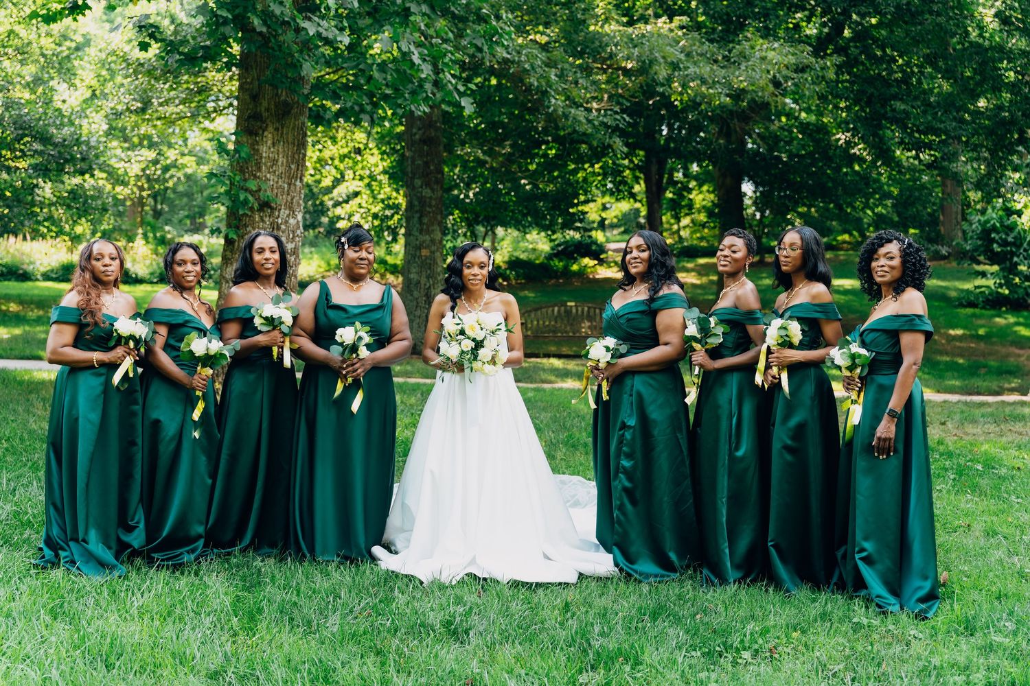 Bride in white gown poses with eight bridesmaids in emerald green off-shoulder dresses holding bouquets outdoors.