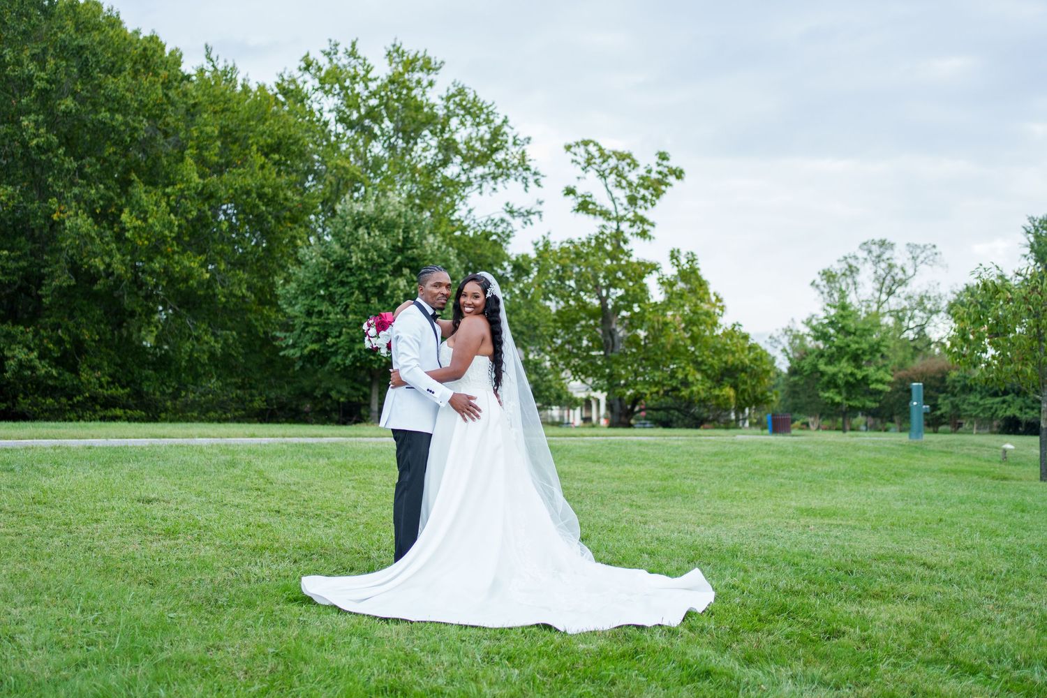 A bride and groom pose together on a lush green lawn surrounded by trees on their wedding day.