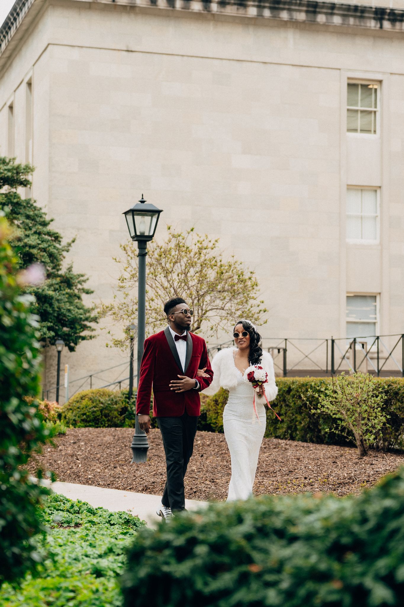 Newlywed couple walks together in garden, groom in burgundy tuxedo, bride in white dress holding bouquet.