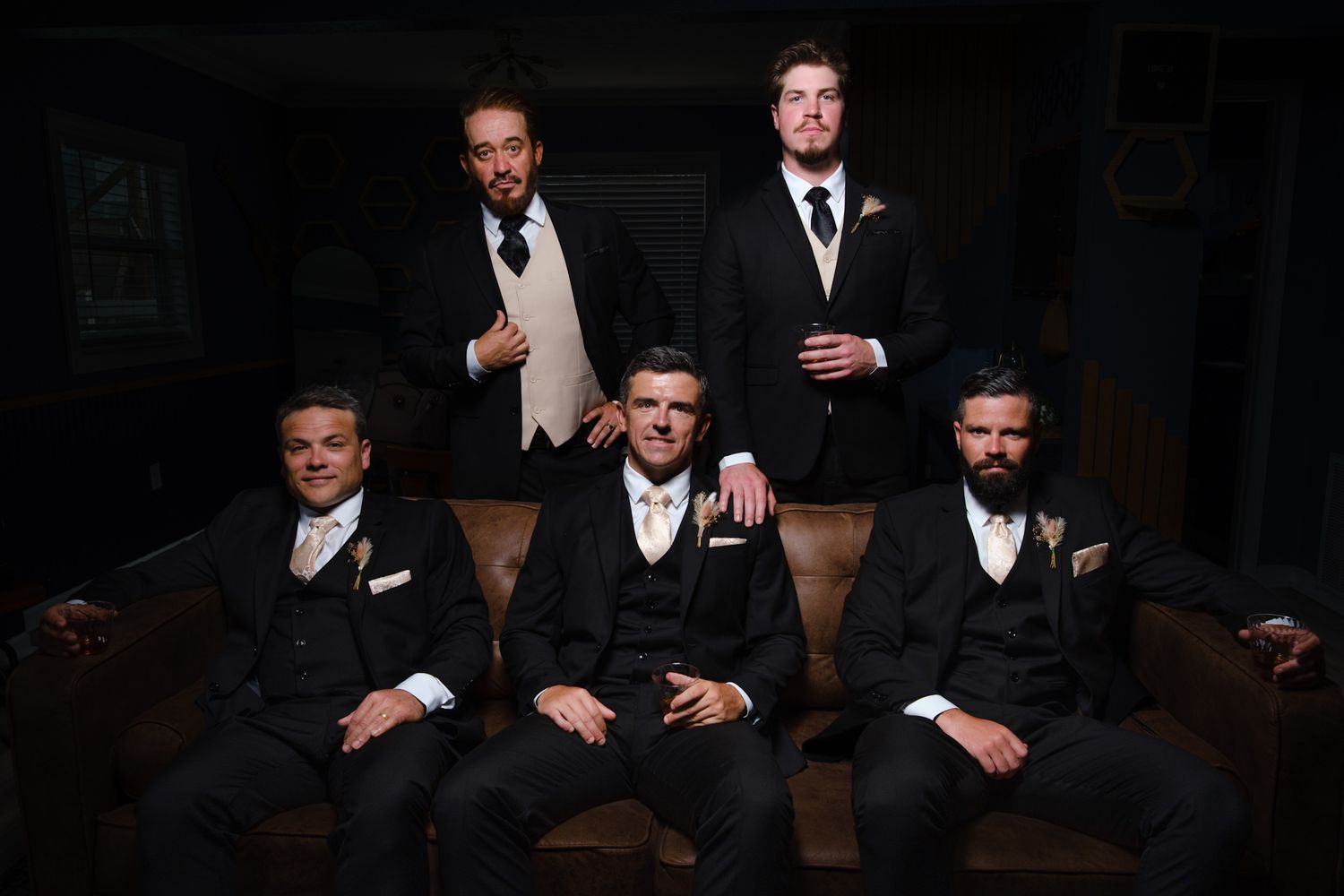 Five groomsmen in formal black tuxedos and bow ties pose together on dark leather furniture in moody lighting.