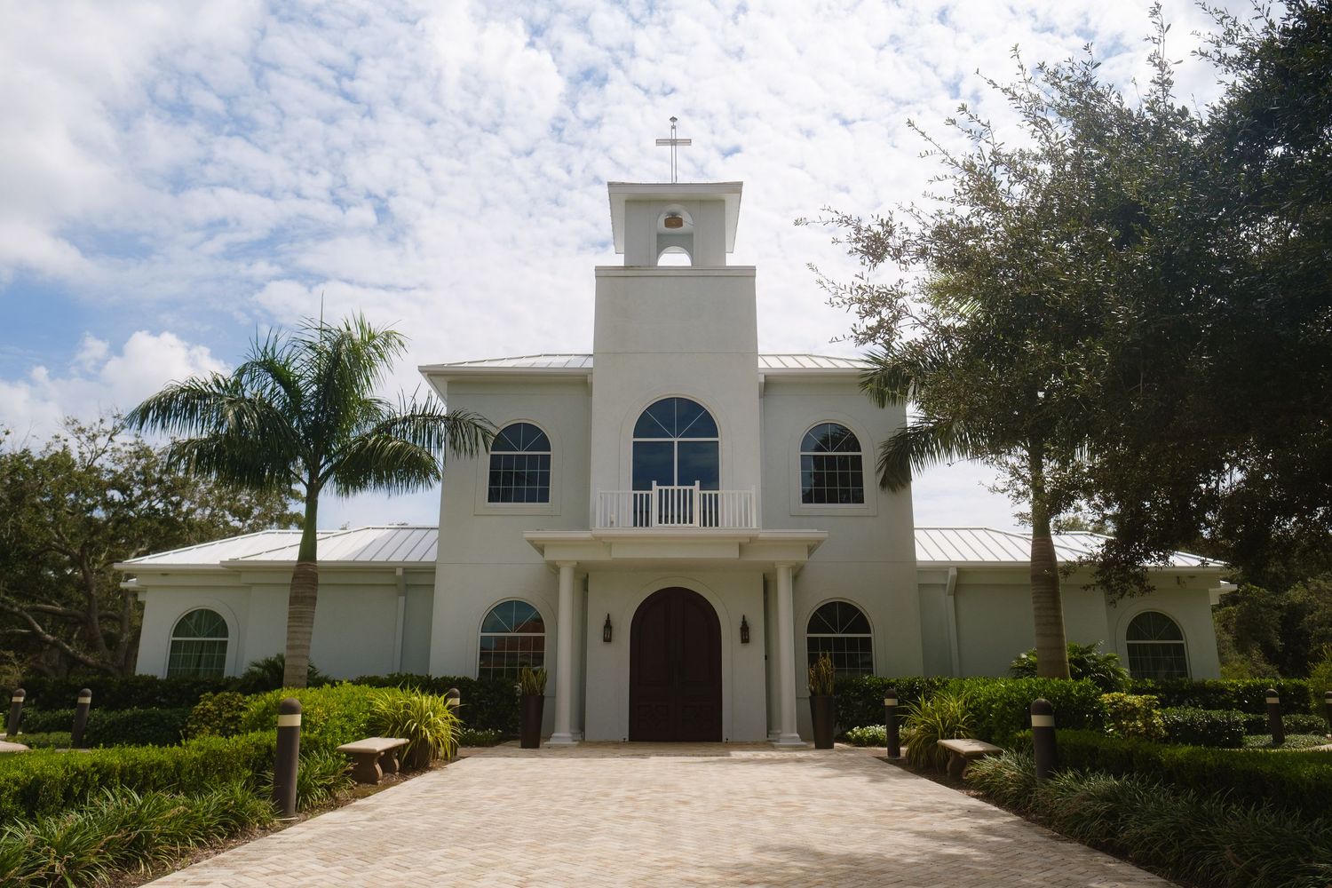 A white stucco church with arched windows and bell tower stands behind manicured hedges and palm trees on a sunny day.