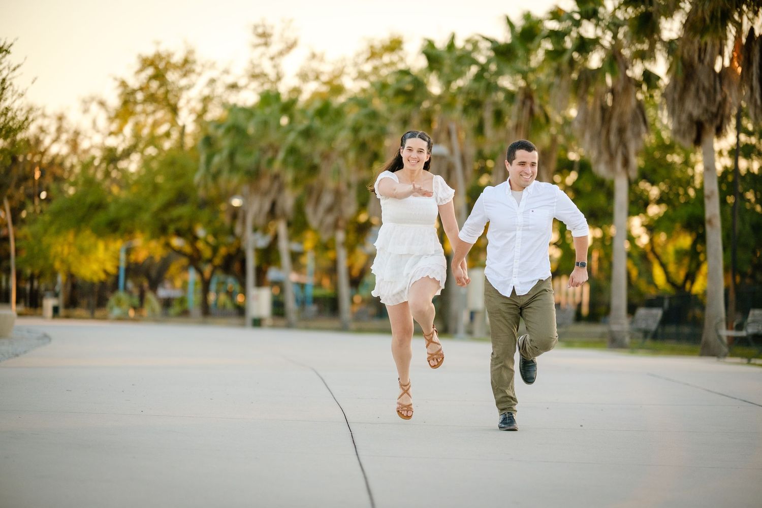 Two people running together on a paved path through a park at sunset with trees in the background.