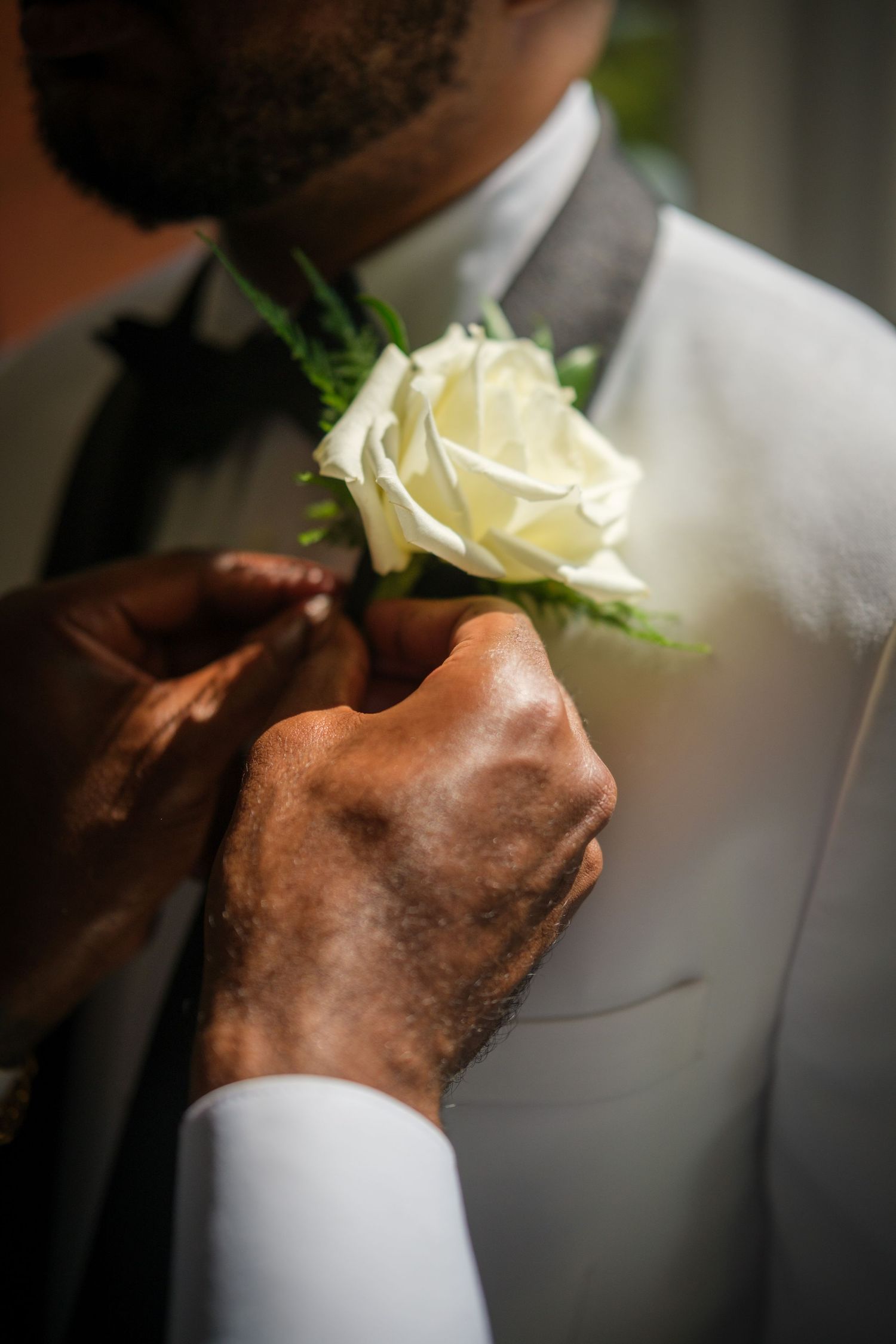 Hands pinning a white rose boutonniere on a groom's white tuxedo lapel at a wedding.