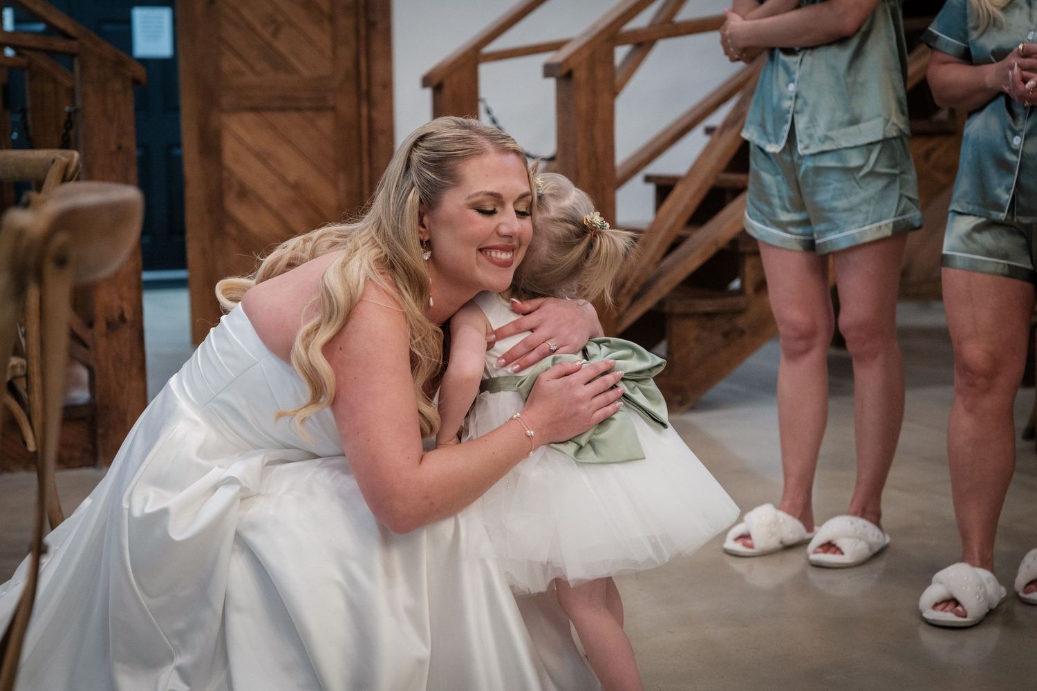 Bride in white gown kneeling to hug a young flower girl in a tutu at a wedding reception.