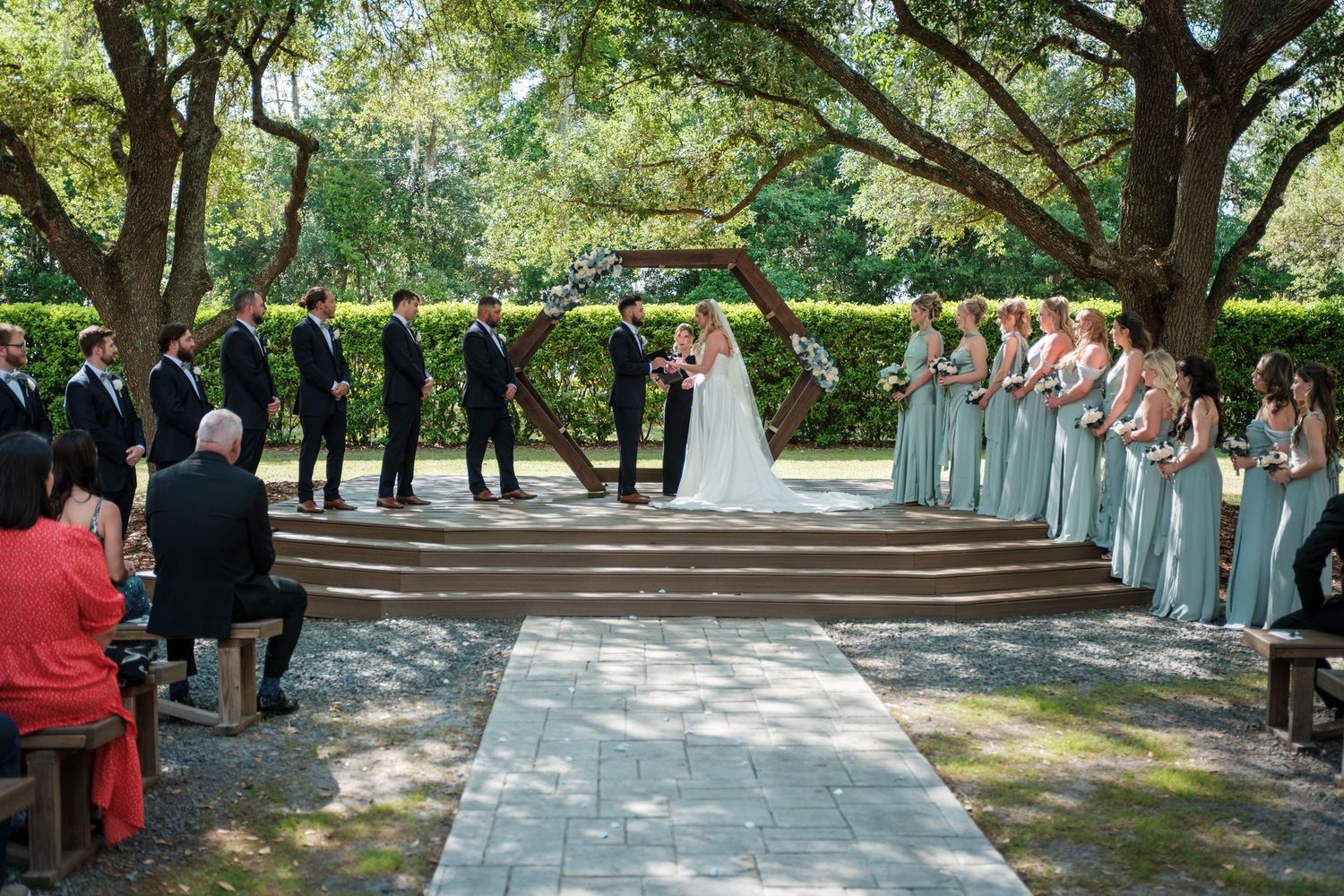 Outdoor wedding ceremony with bridesmaids in sage green dresses, groomsmen in black suits, under lush trees on a stone path.